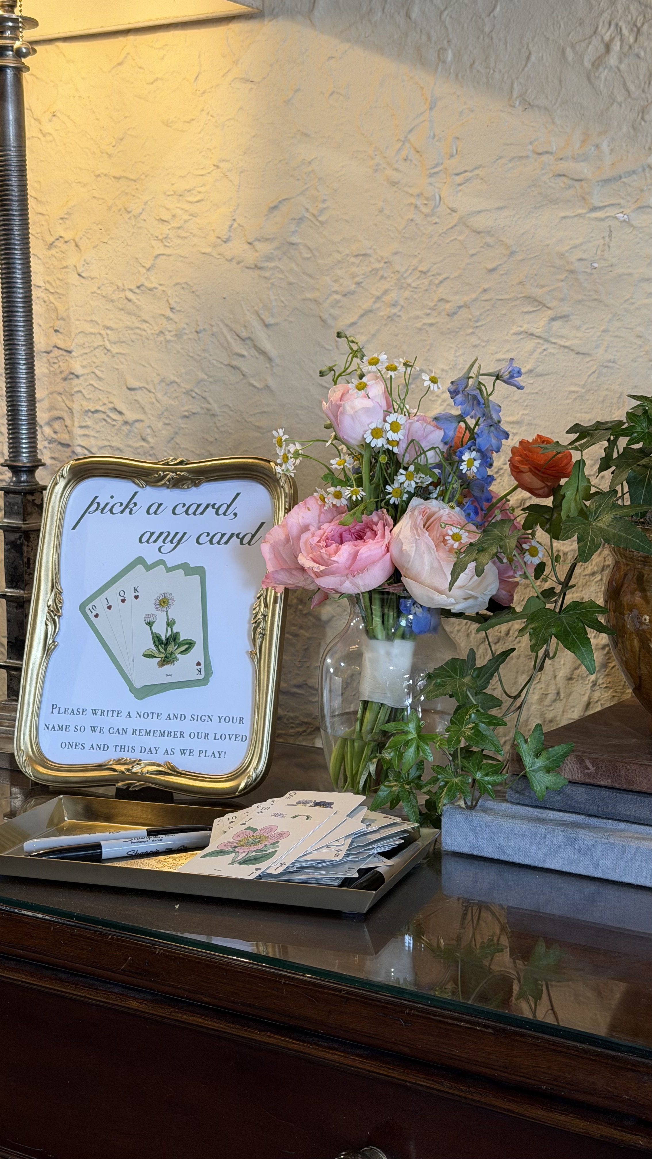 A sign instructs visitors to pick a card and write a note, with a small table of playing cards, pens, and a tray nearby. Beside the sign, there's a vase of mixed pink, white, and blue flowers, surrounded by green ivy, on a wooden table against a text