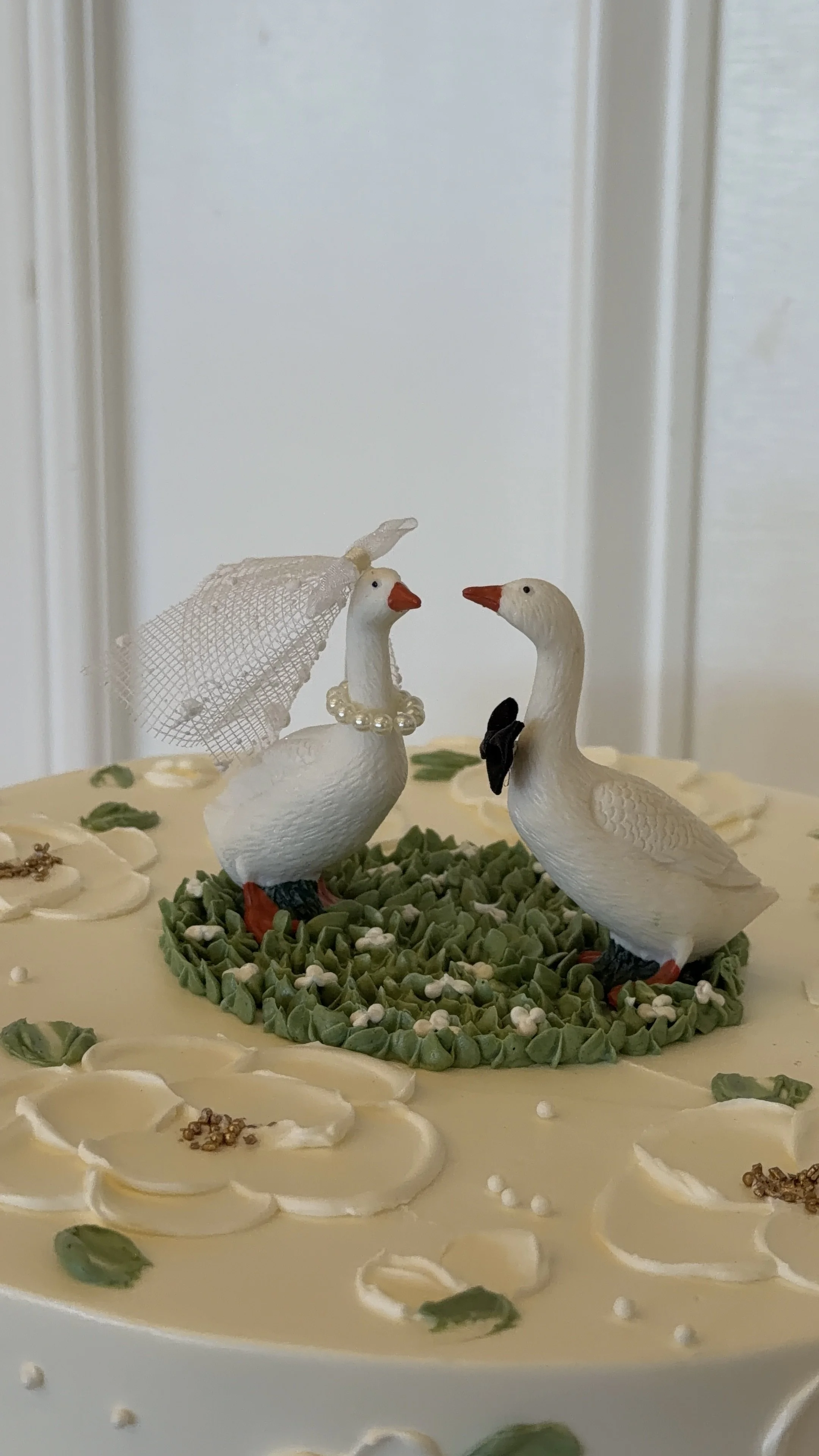 Decorative cake with two white duck figurines, one wearing a veil and pearl necklace, the other with a black bow, on a green grassy base surrounded by white and green icing flowers.