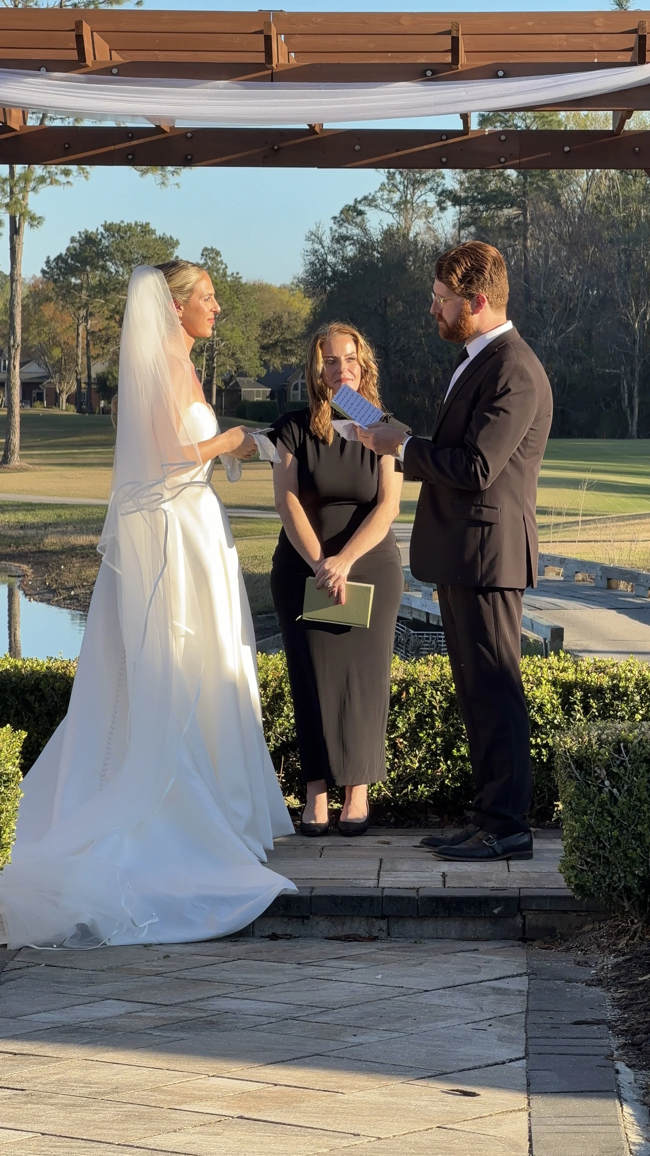 A wedding ceremony taking place outdoors with a bride in a white wedding gown and veil, a groom in a black tuxedo, and an officiant woman in a black dress. They are standing on a small wooden platform with greenery around, and a scenic background of 