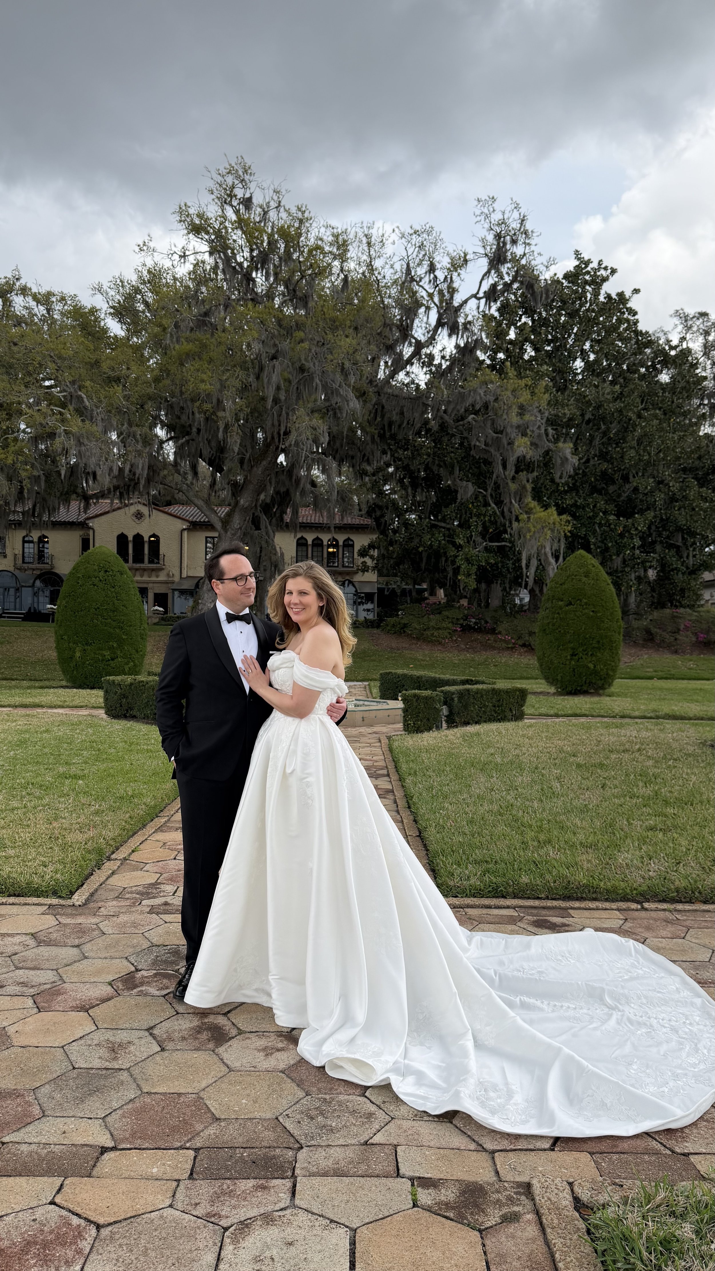A bride and groom standing together outdoors on a paved path, with a large tree and grand building in the background during overcast weather.