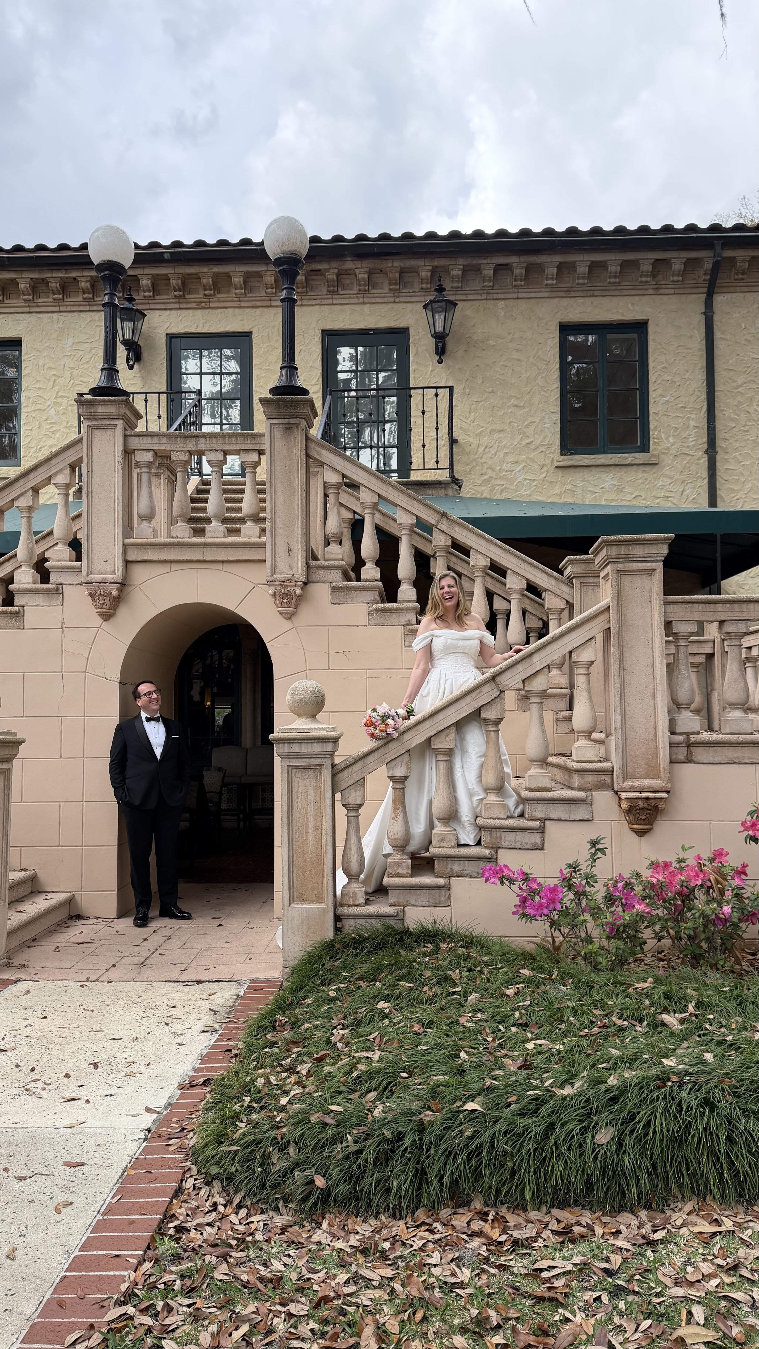 A bride in a white wedding dress standing on a stone staircase, holding a bouquet of flowers, smiling, with a groom in a black tuxedo standing below in front of a brick archway, outside a vintage-style building with yellow walls and green shutters.