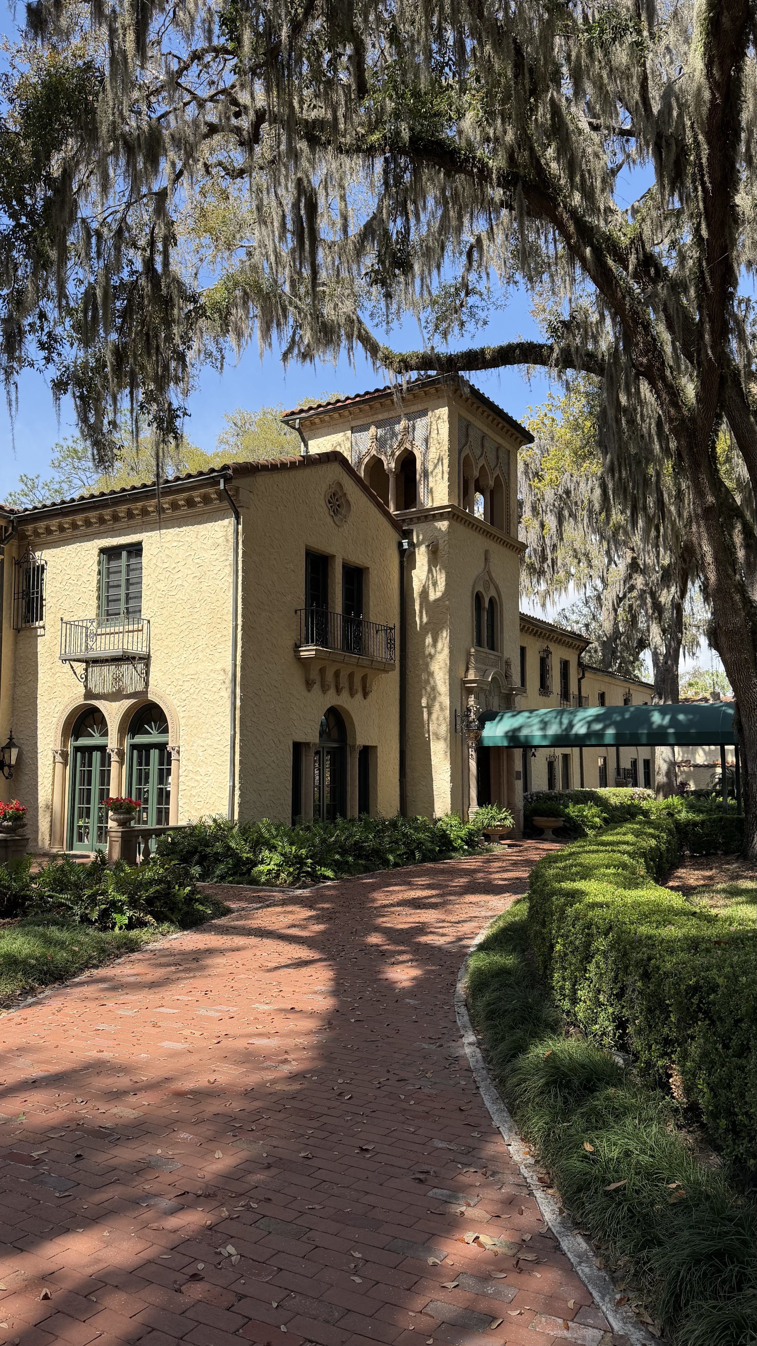 A large, old-fashioned house with a Spanish-style architecture featuring arched windows and a tower, surrounded by trees and a brick pathway, under a bright blue sky.