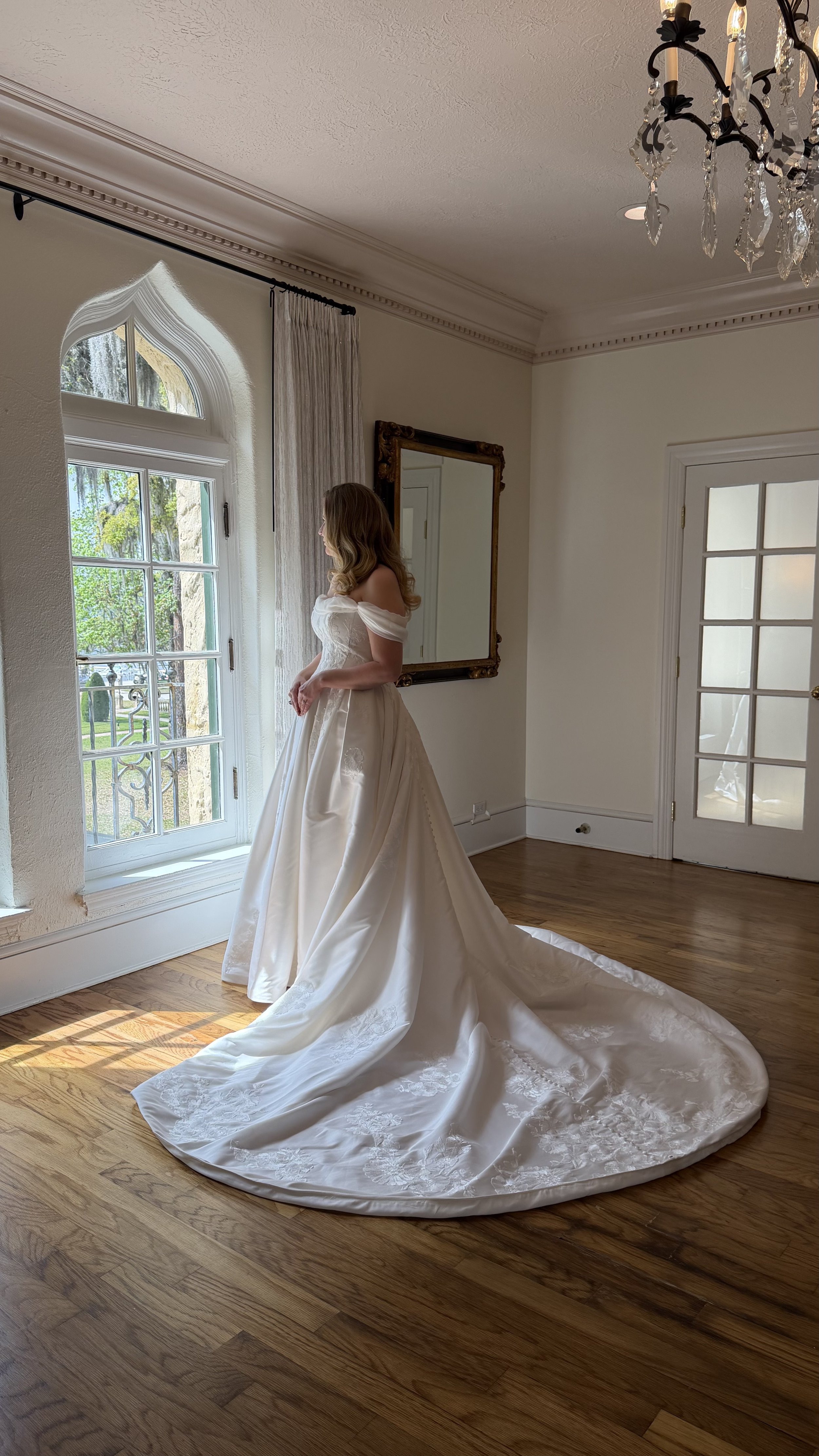 A woman in a wedding dress standing near a window in a room with wooden floors.