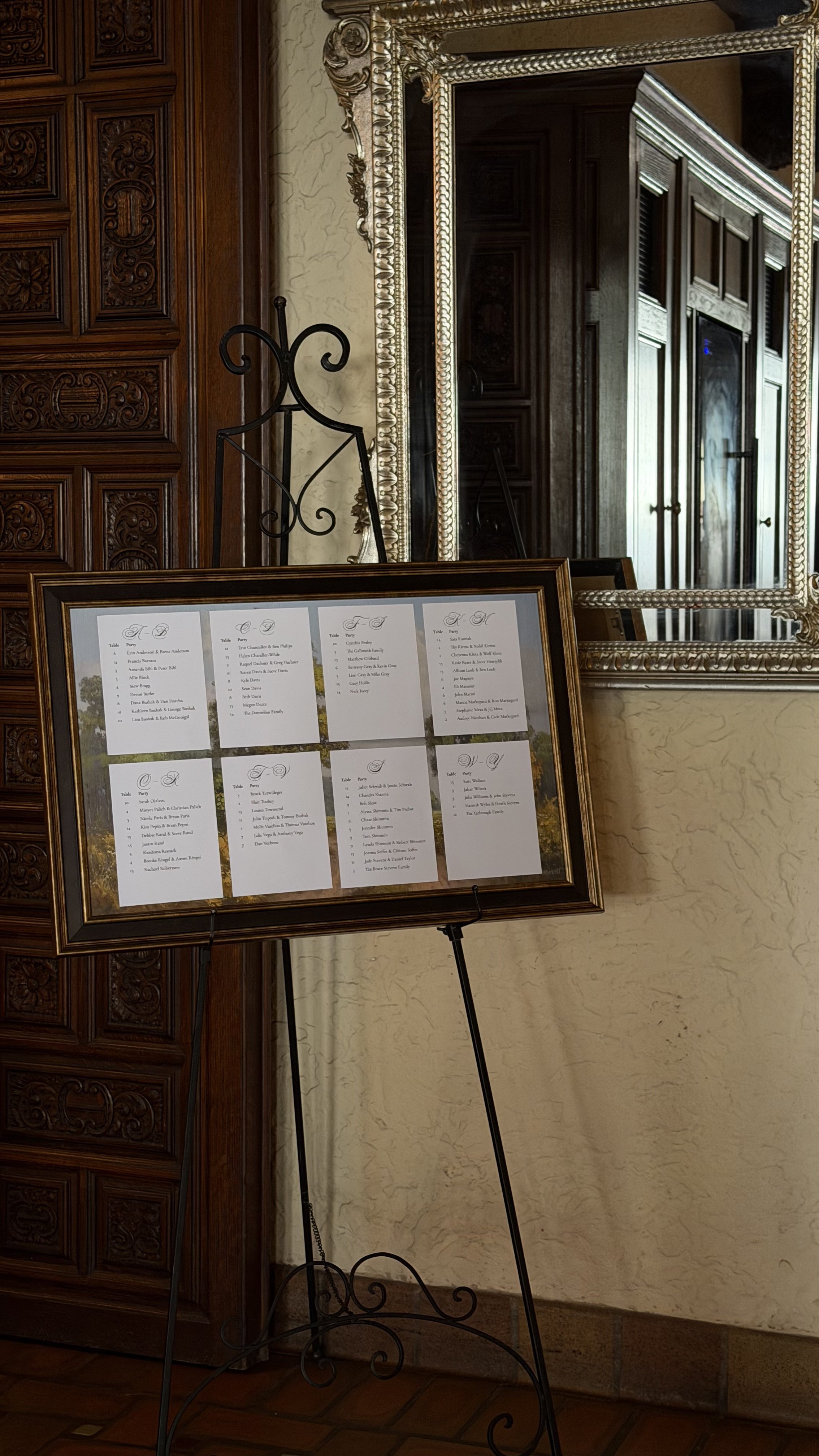 A framed seating chart for a wedding or event, displayed on a black metal stand next to a large ornate mirror and wooden wall paneling.
