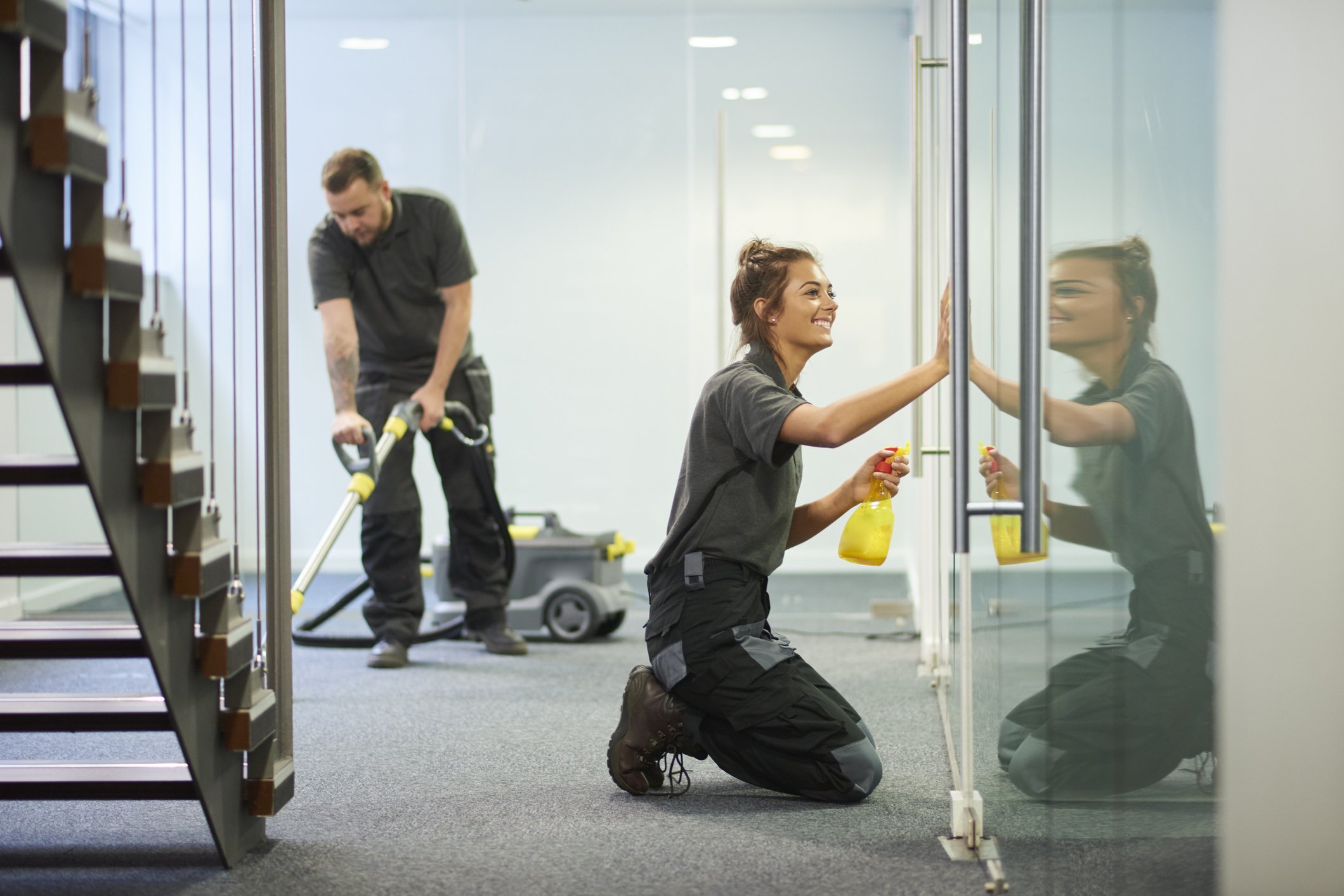 Two cleaning workers, a woman kneeling and a man standing, cleaning glass doors with spray bottles and a vacuum cleaner in a modern office space.