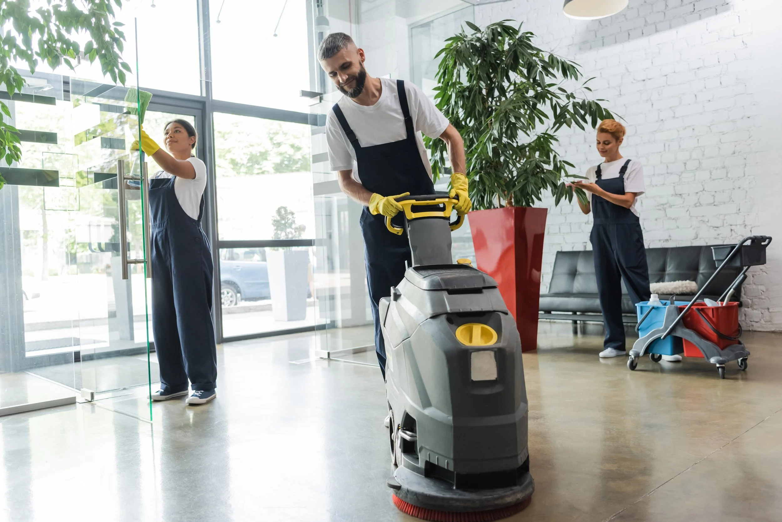 Cleaning staff cleaning the lobby of a building with a floor scrubber, glass door, and cleaning supplies.