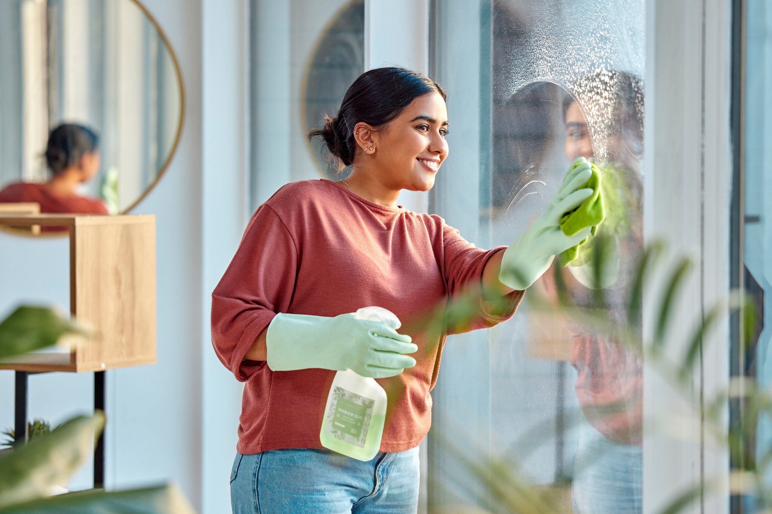 A young woman in a red shirt and jeans cleaning a glass window with a spray bottle and a green cloth, smiling.