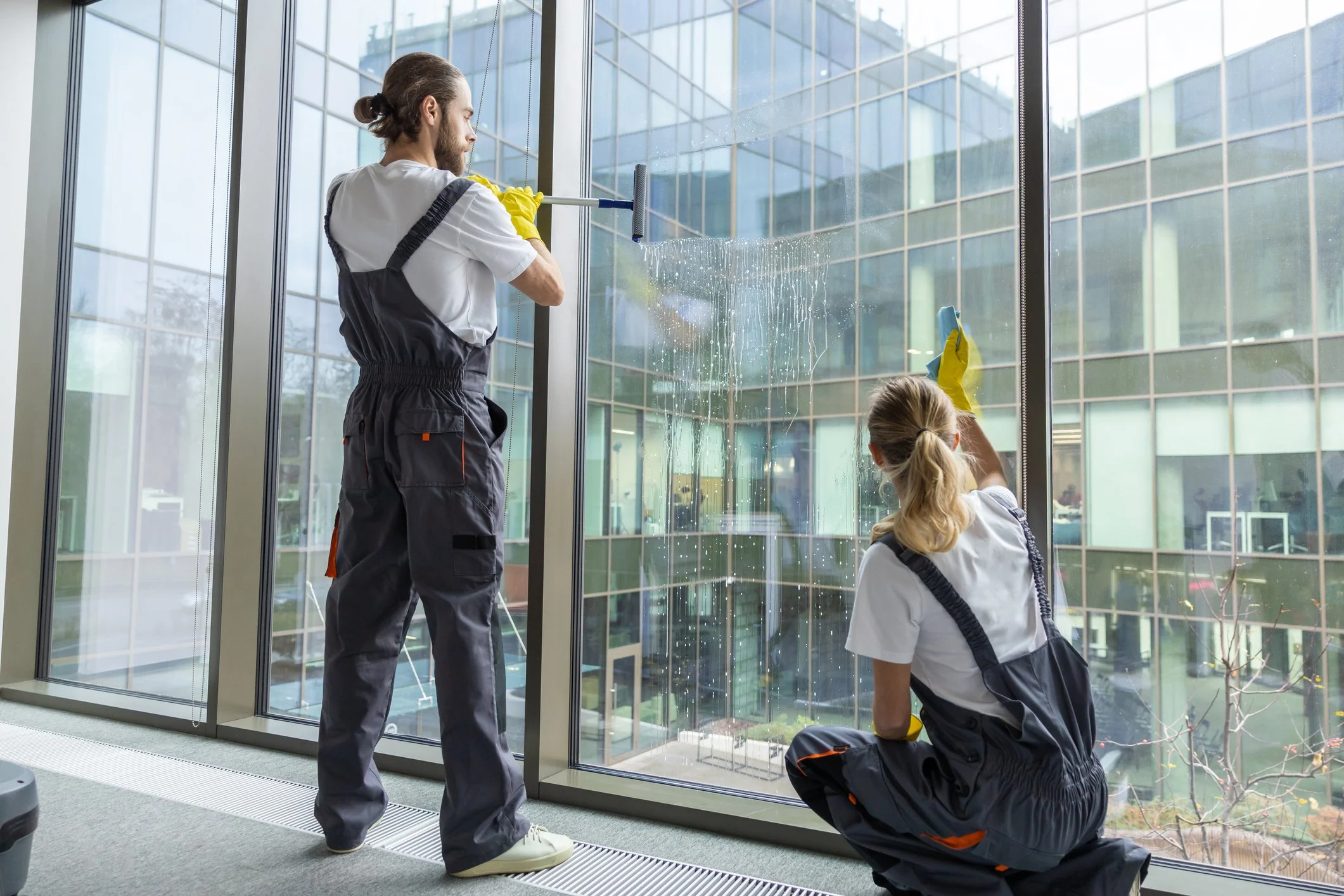 Two window cleaners, one standing and one kneeling, cleaning large glass windows of a modern building with water and squeegees.