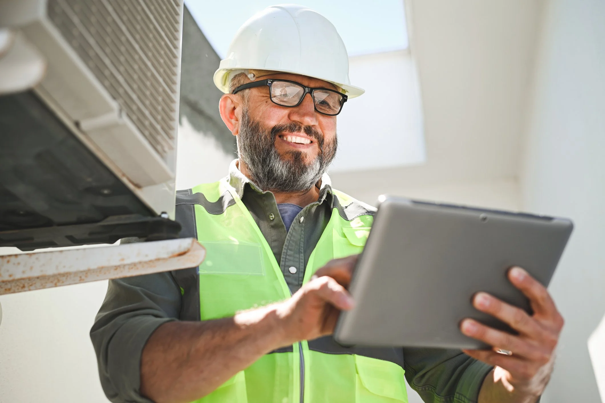 A smiling construction worker with a beard, glasses, and a white hard hat, wearing a neon yellow safety vest, using a tablet in a bright, indoor setting.