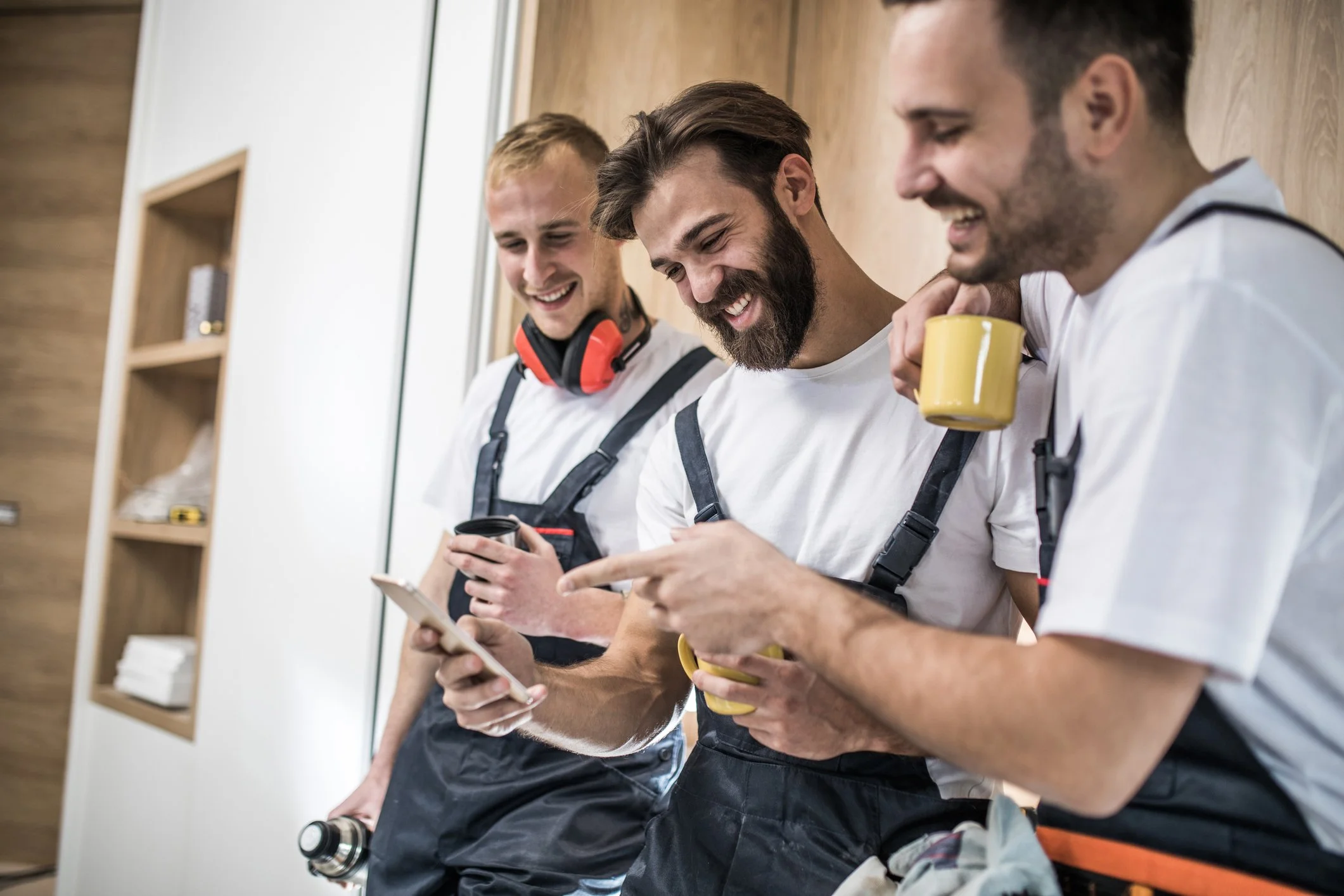 Three men in work overalls standing in a room, smiling and looking at a smartphone together. One of them is holding a yellow mug and another has a coffee cup.