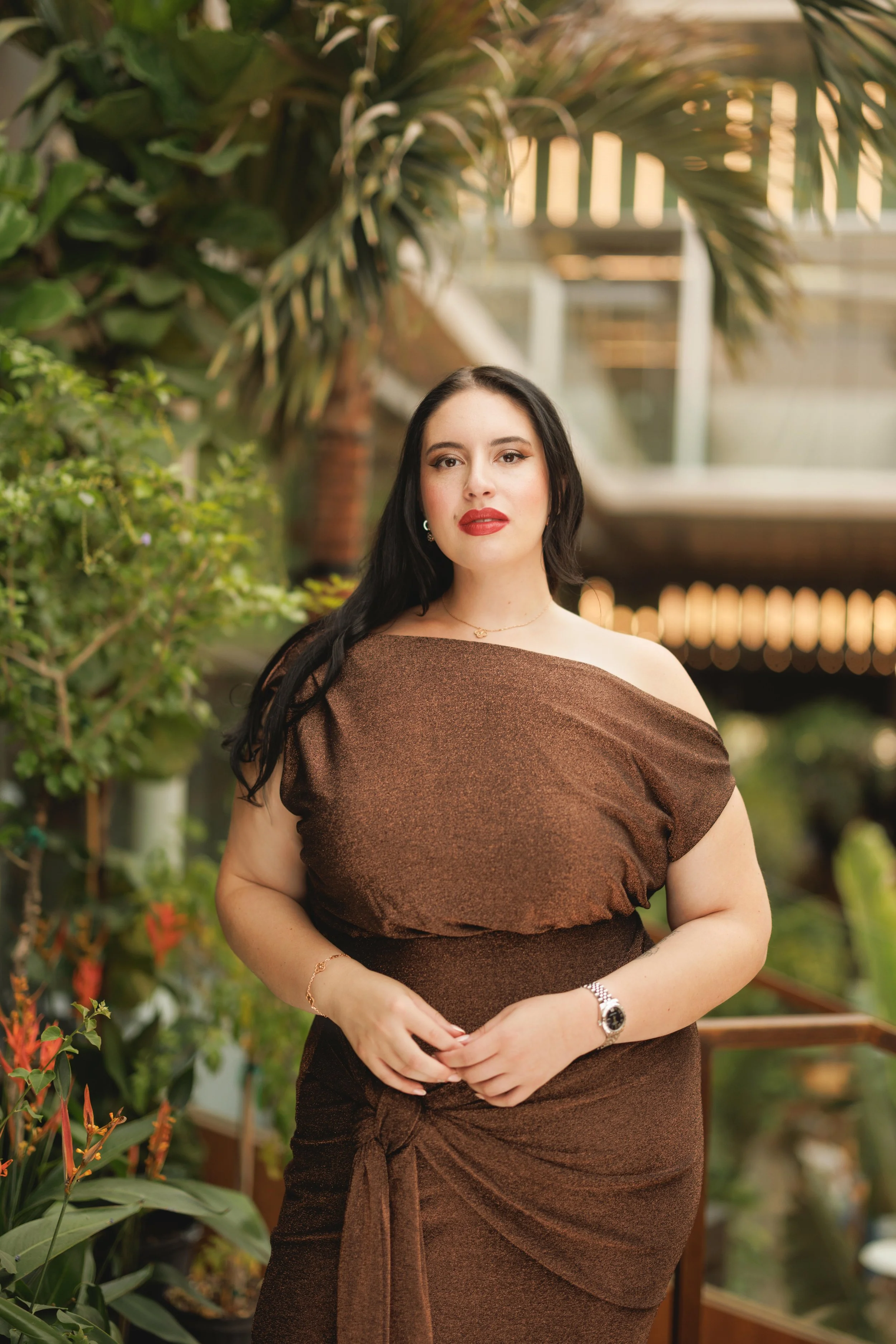 A woman with long black hair and red lipstick standing in front of green plants and a modern building in an outdoor setting.