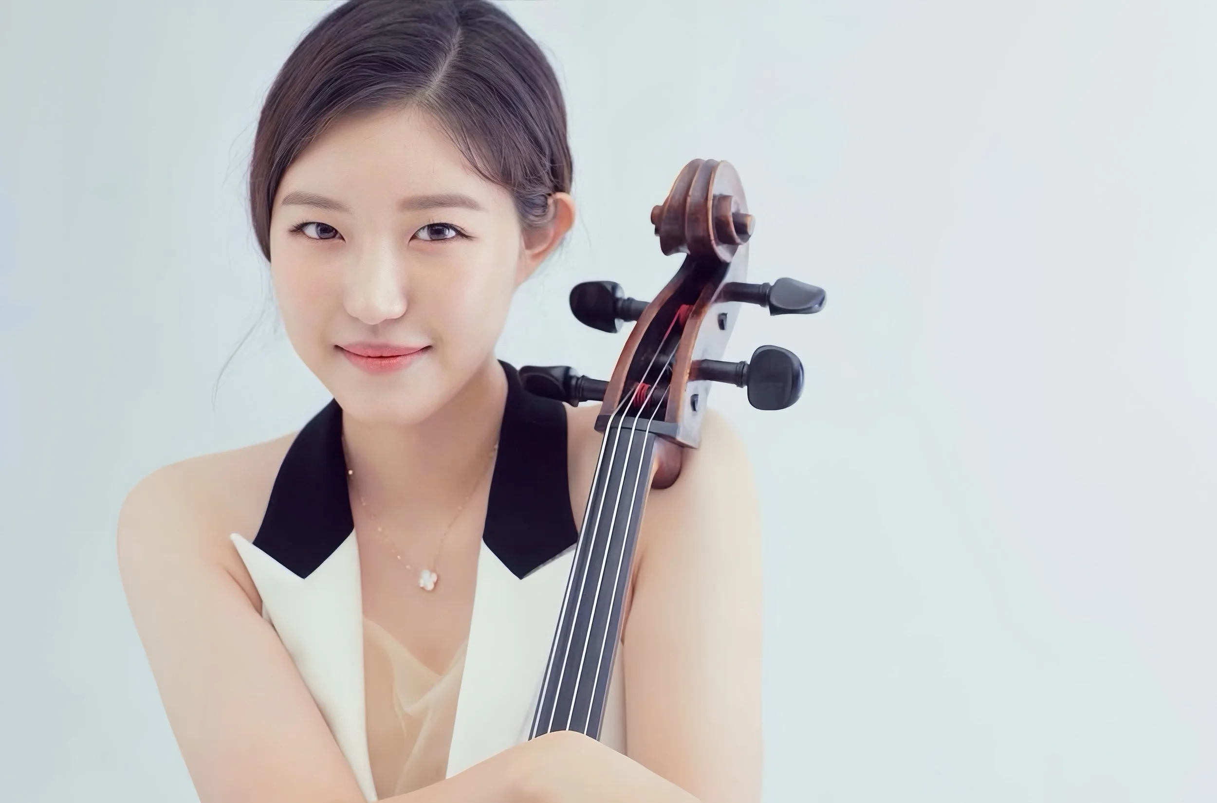 A young woman with short dark hair holding a cello, smiling, against a plain light-colored background.