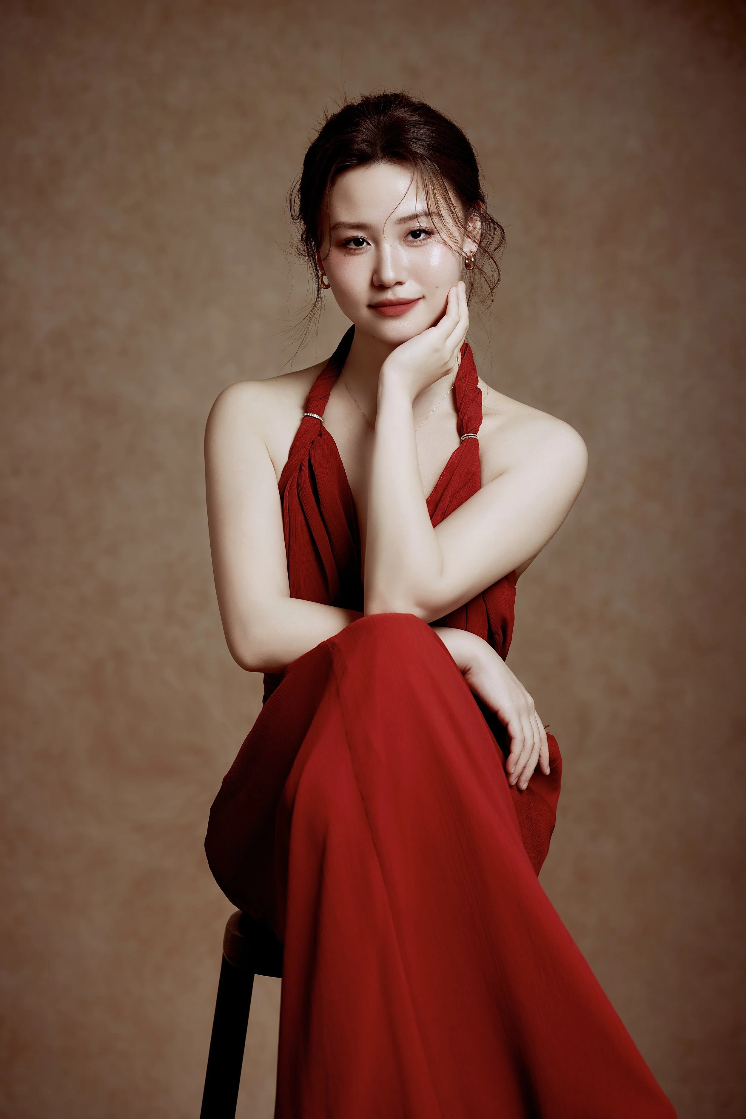 A woman with dark hair styled in a loose updo, wearing a red halter dress, sitting on a stool against a plain brown background, resting her chin on her left hand and smiling softly.