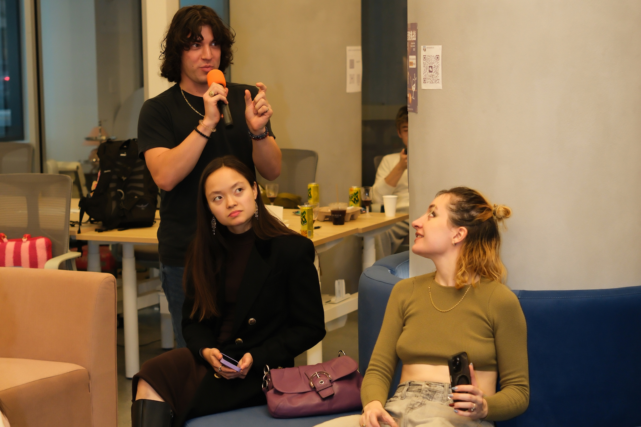 A young man with curly hair holding a microphone and speaking to two young women seated on a blue and green couch in a casual indoor setting.