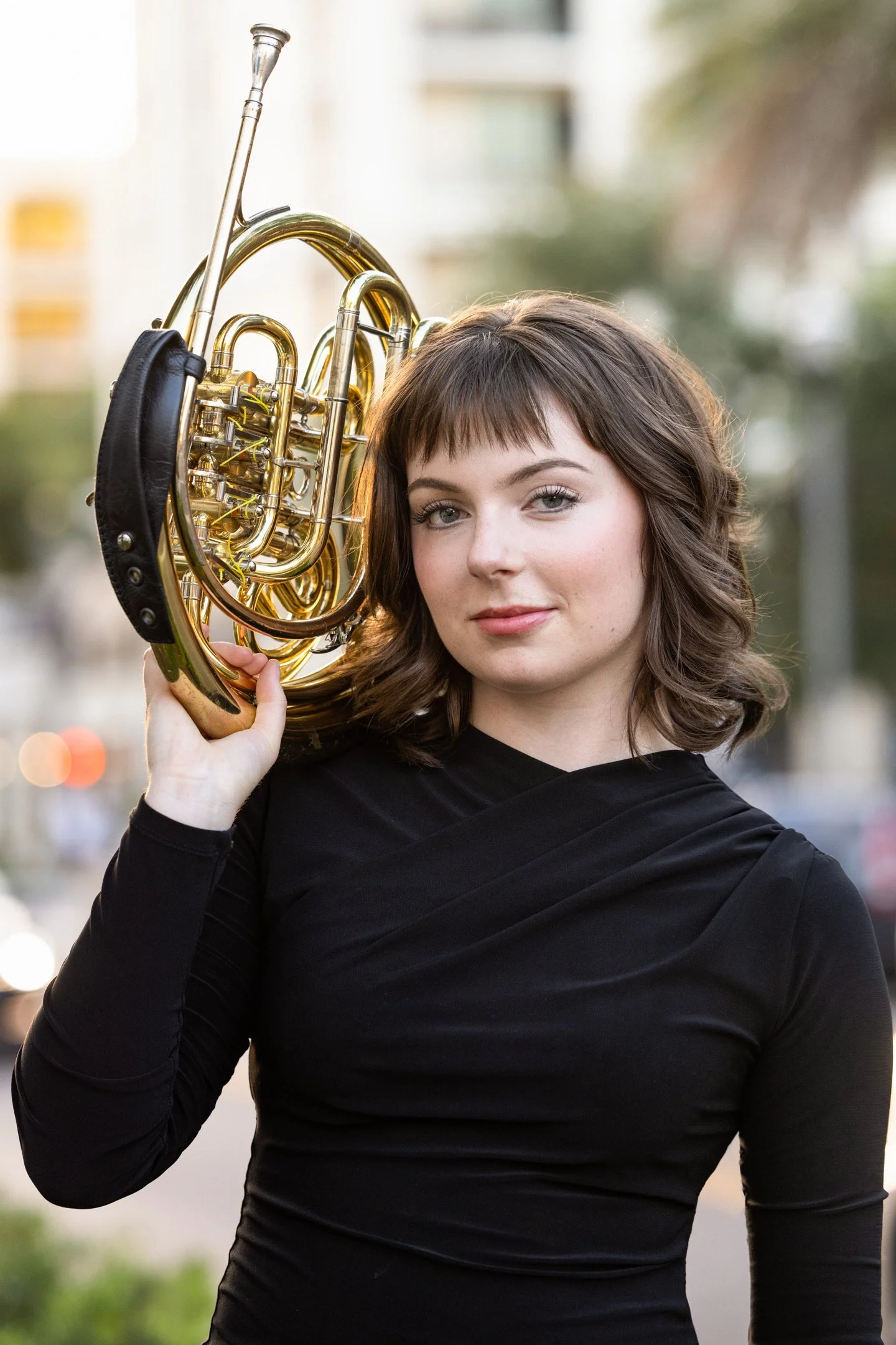 A young woman with shoulder-length brown hair holding a brass French horn on her shoulder outdoors. She is wearing a black long-sleeve top, and the background is blurred with trees and buildings.