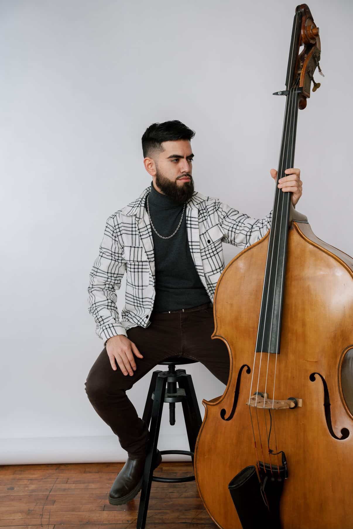 A man with dark hair and a beard sitting on a black stool, playing a large wooden double bass against a plain white background.