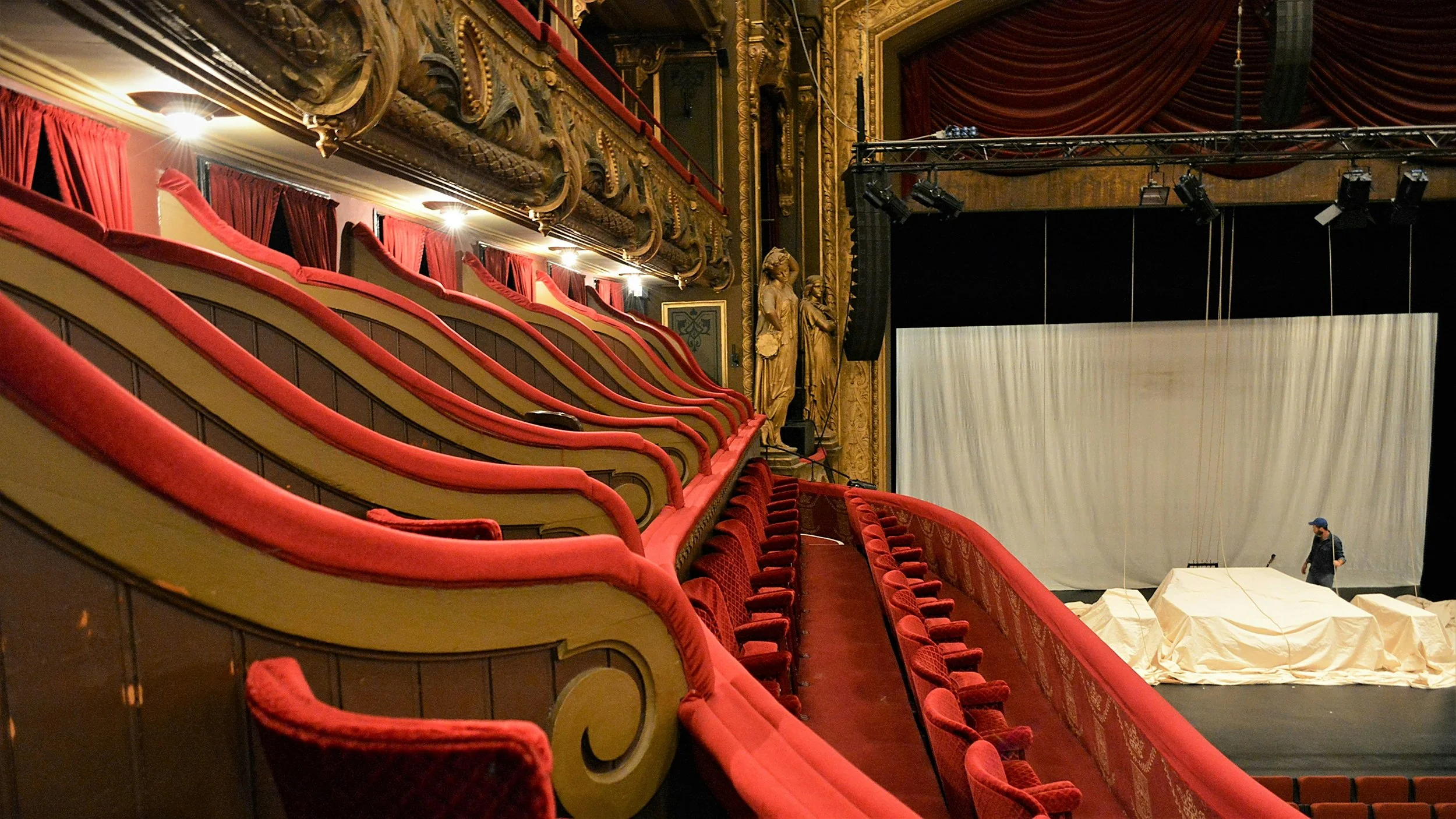 Empty theater with red velvet seats and ornate gold decor, stage with white backdrop, and a person near the stage setting up.