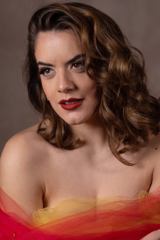 A woman with shoulder-length wavy brown hair, wearing red lipstick and eye makeup, gazes slightly to the side. She is draped in sheer, colorful fabric around her shoulders and chest, with a plain brown background.