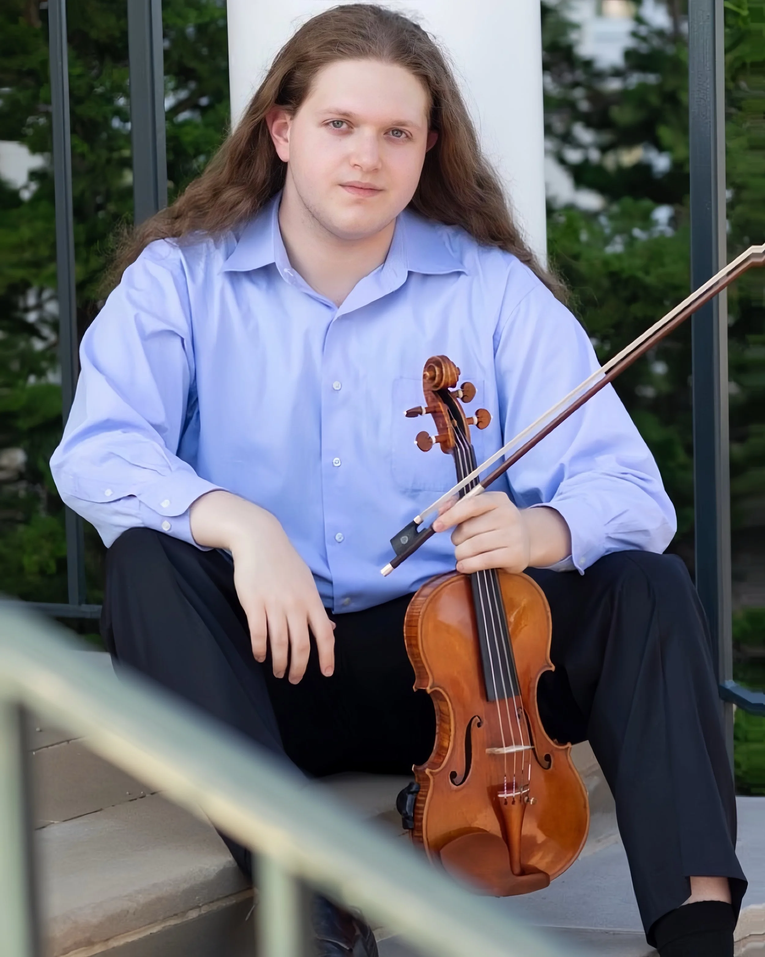 Young man with long hair sitting on stairs outdoors, holding a violin and a bow, wearing a light blue shirt and black pants.
