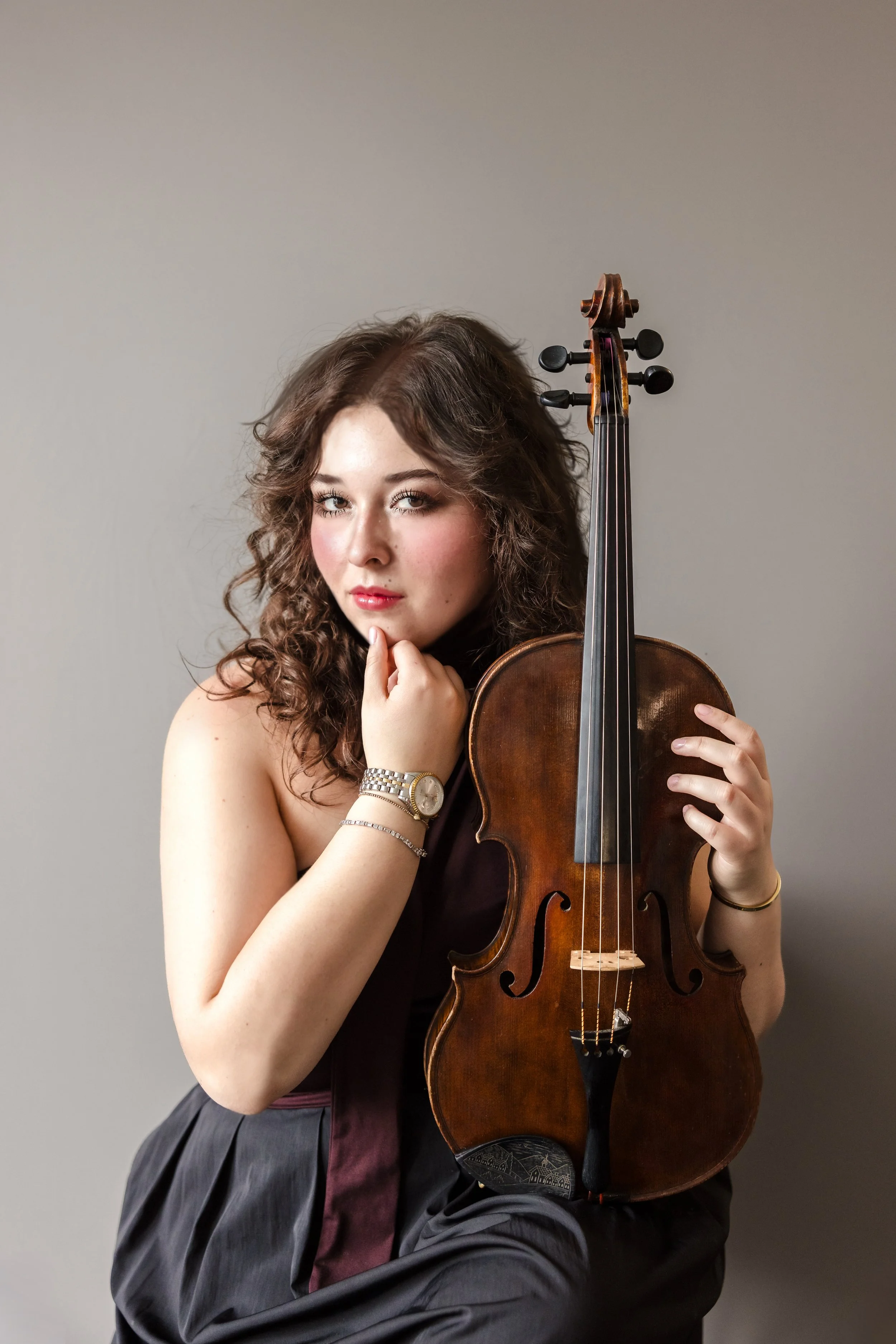 A young woman with curly brown hair holding a violin close to her chest, posing against a plain background.