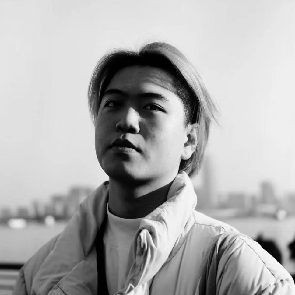 Black and white portrait of a young person with light hair, wearing a light jacket, standing outdoors with a cityscape and water in the background.