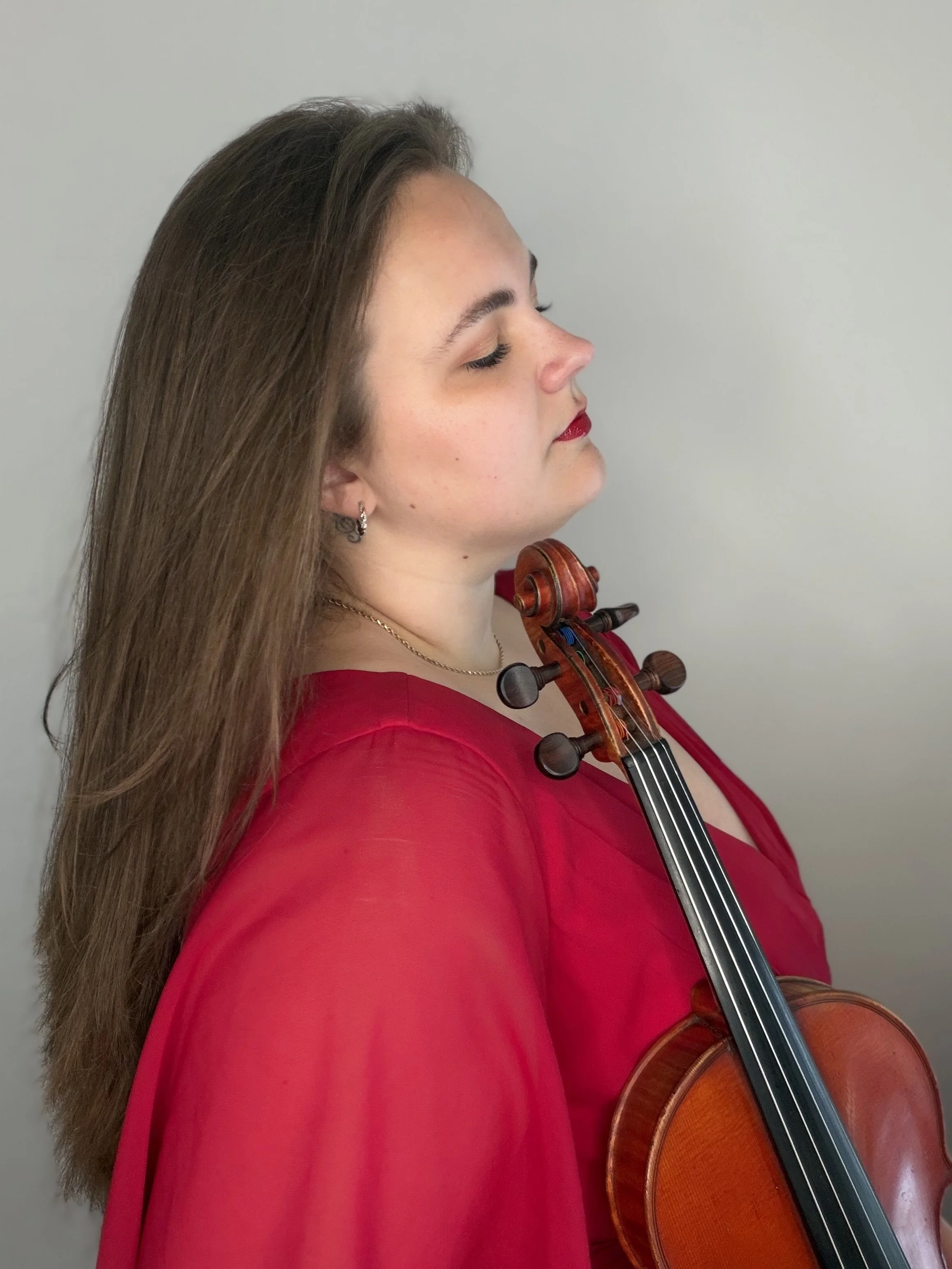A woman with long brown hair wearing a red blouse, holding a violin, with her eyes closed and a serene expression.