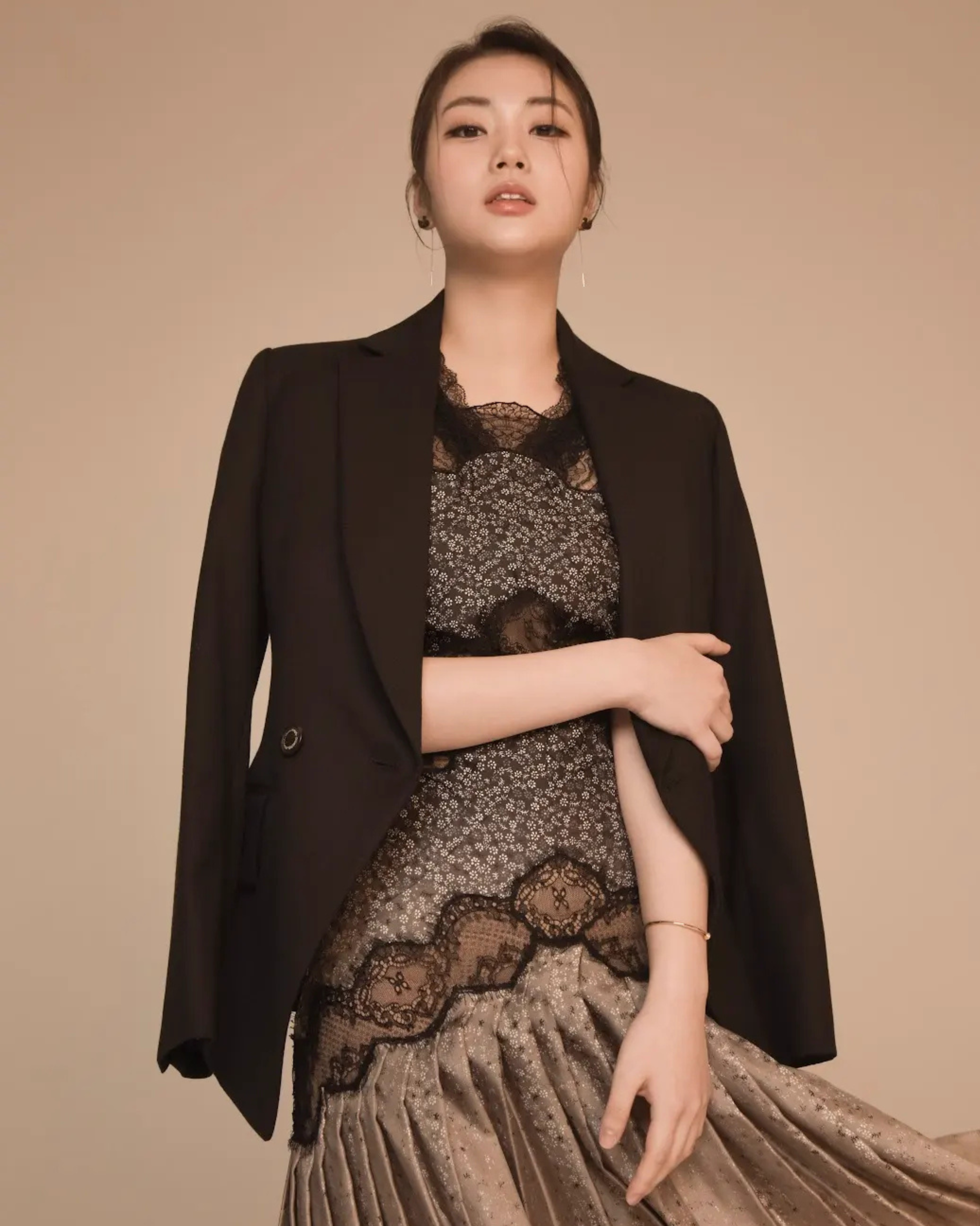 A woman wearing a black blazer over a lace dress, posing with one arm across her waist and the other hanging down.