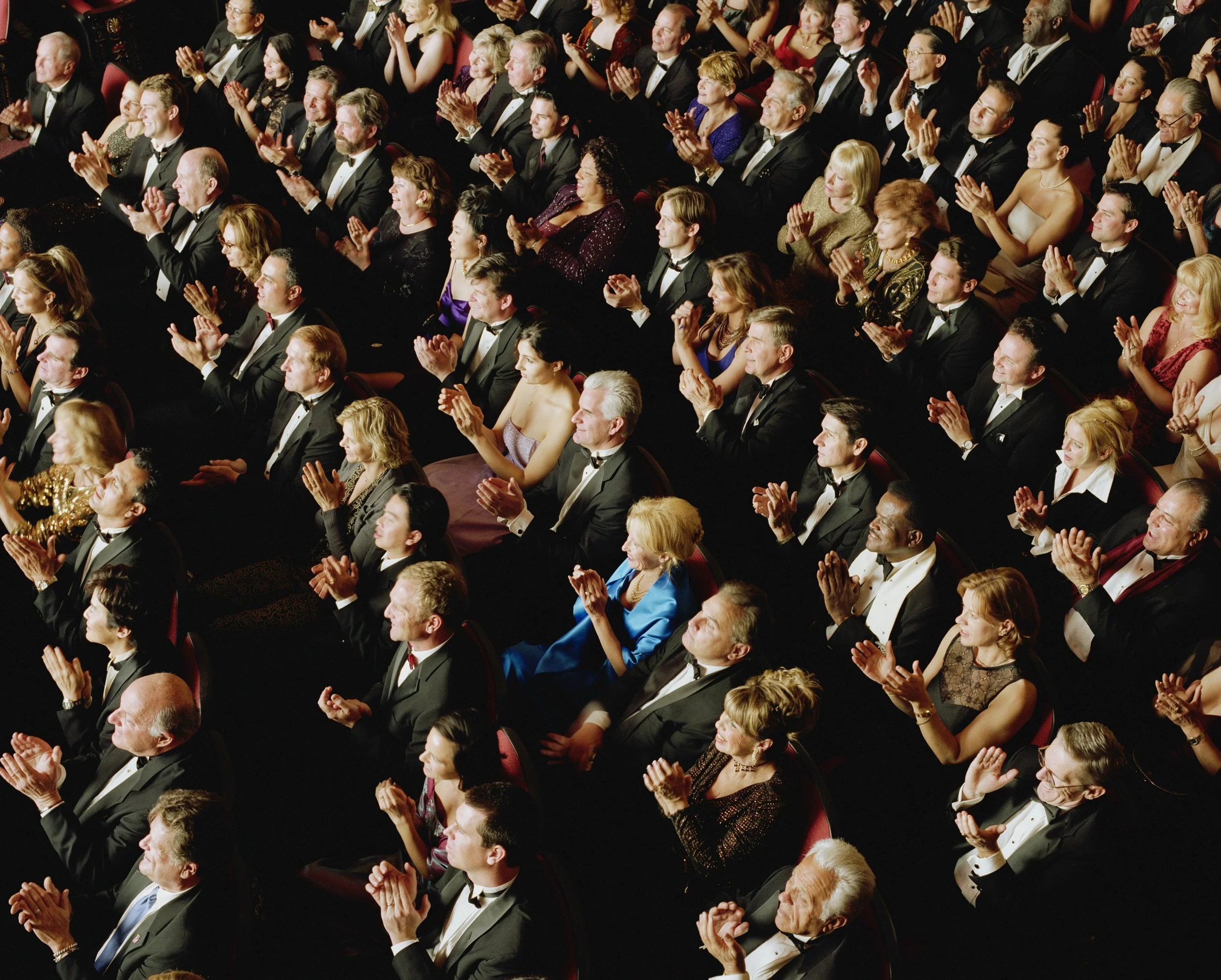 Audience members dressed in formal attire, including tuxedos and evening gowns, sitting in theater seats and applauding during a performance or event.