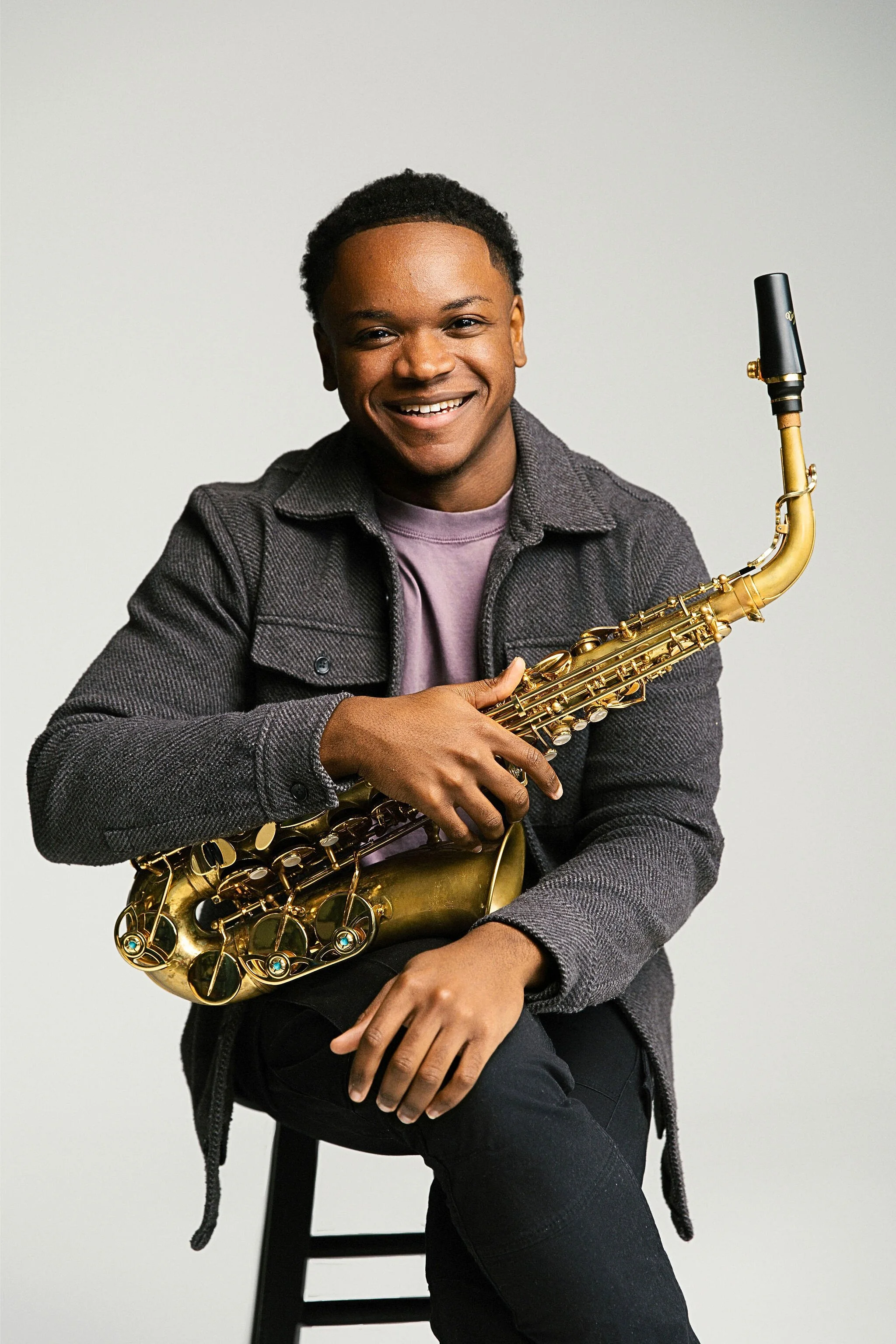 A young man smiling while holding a saxophone, sitting on a stool against a plain white background.