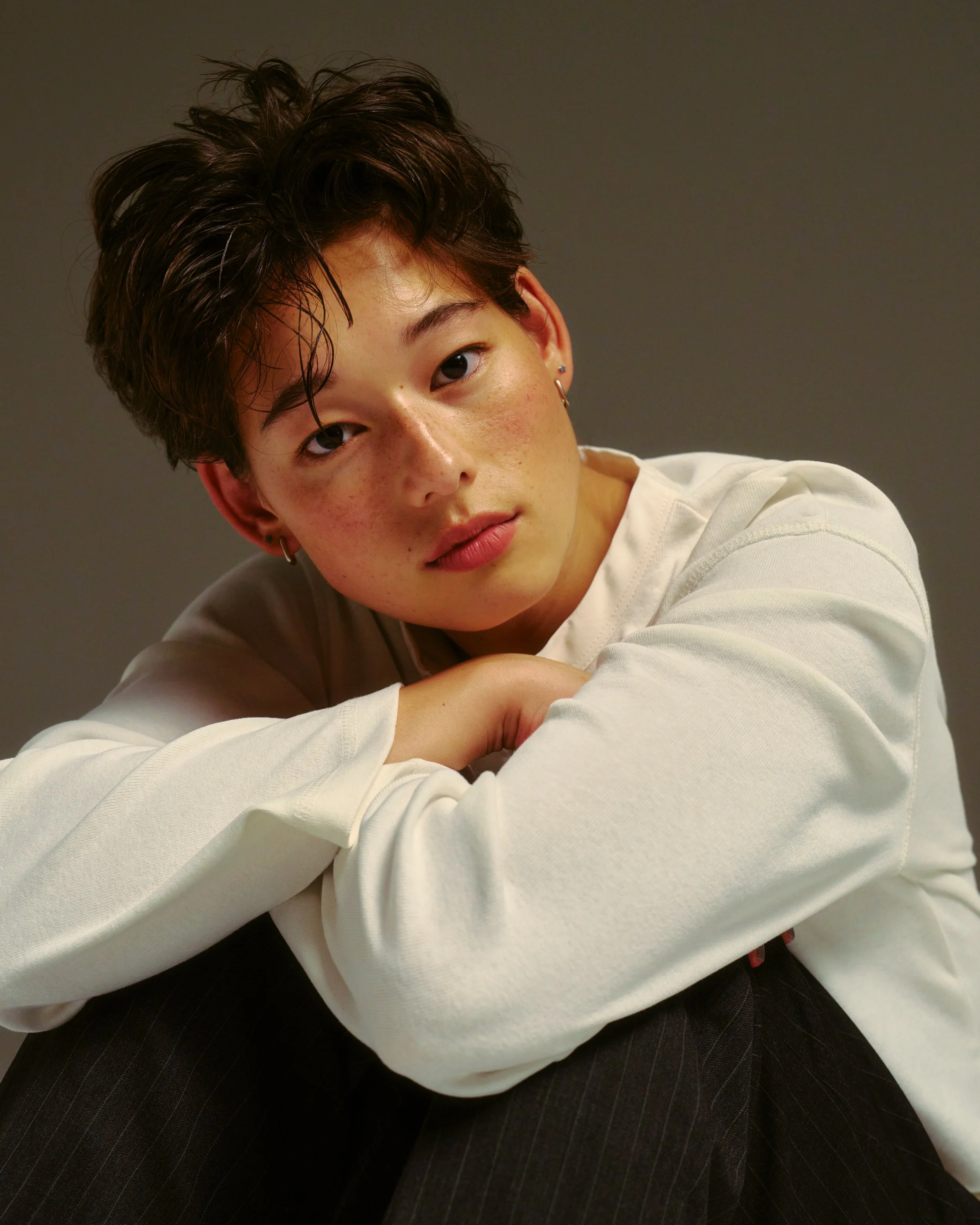 A young woman with short, wavy hair and earrings, wearing a white long-sleeve shirt, resting her arm on her knee and looking at the camera against a dark gray background.