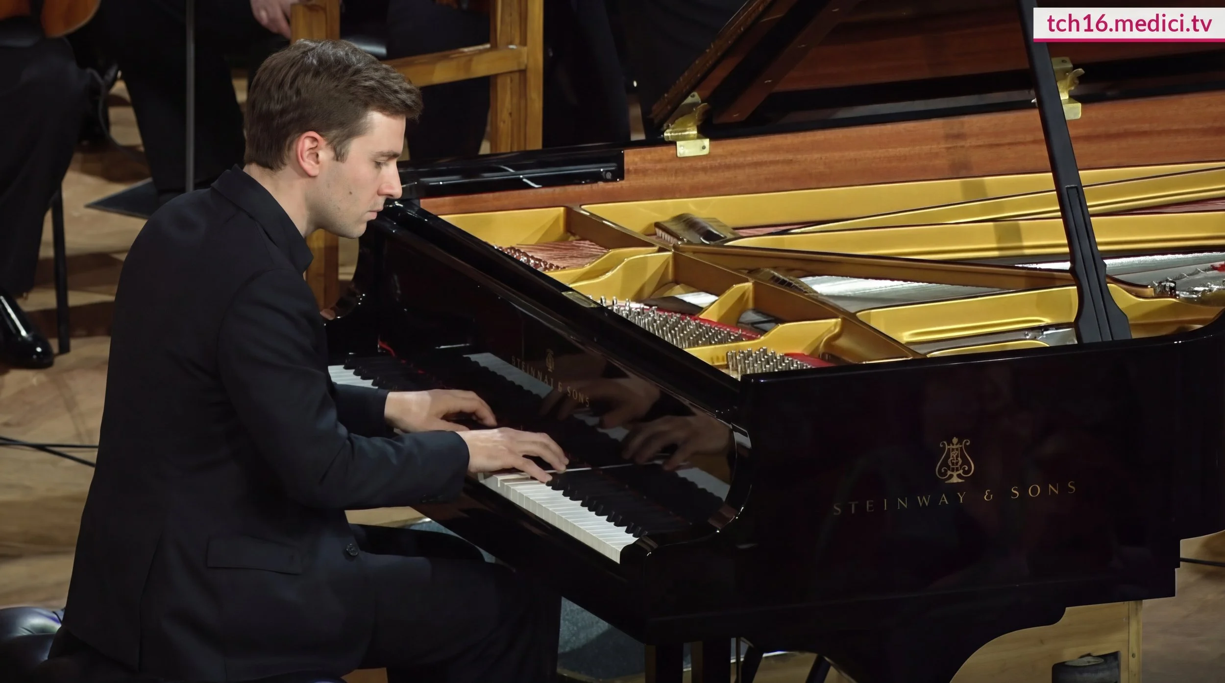 A grand piano in a large, empty concert hall with wooden floors and ornate decor.