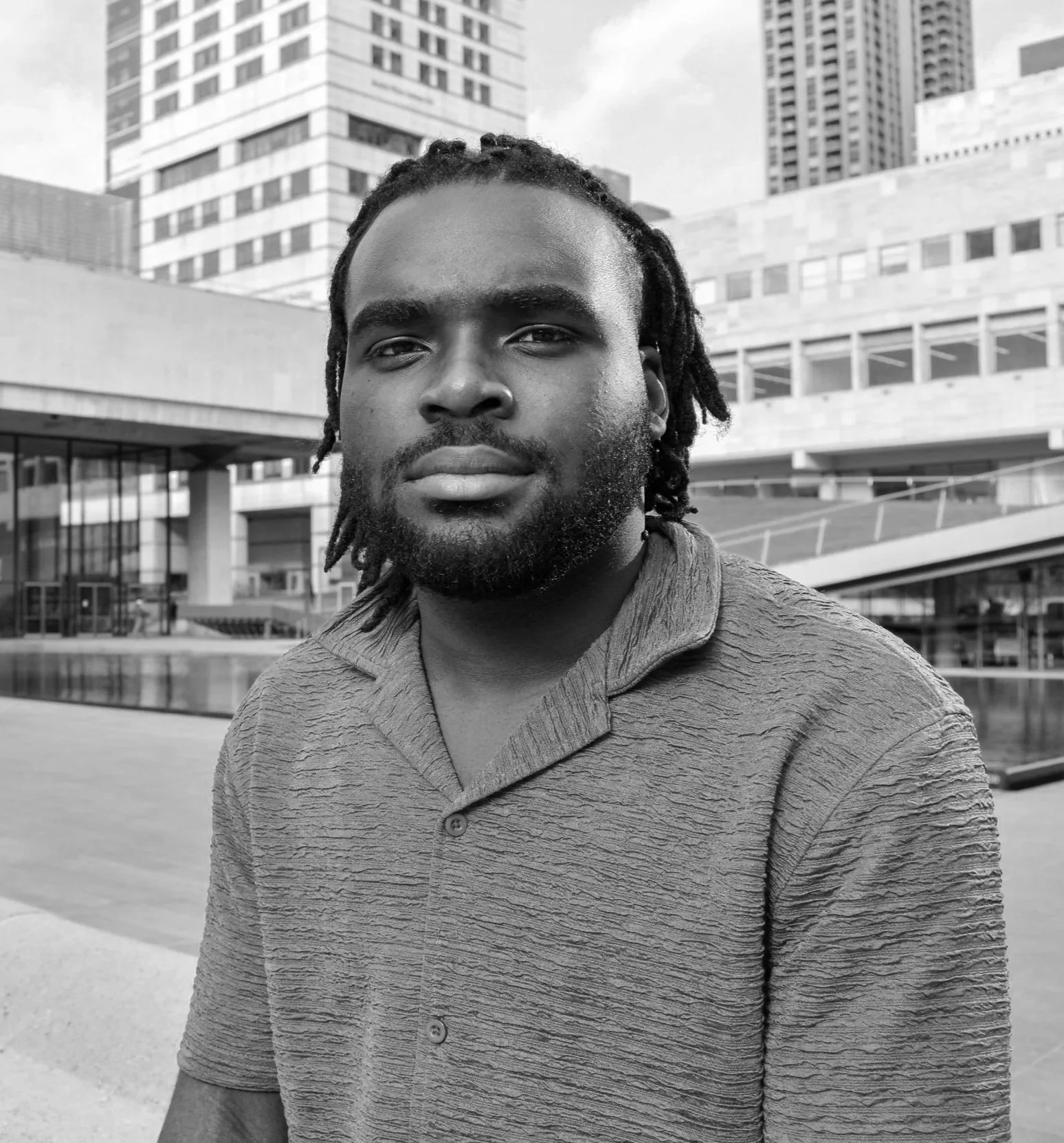 A black and white photo of a man with dreadlocks, beard, and mustache, wearing a textured short-sleeve shirt, standing outdoors in front of modern buildings in an urban setting.