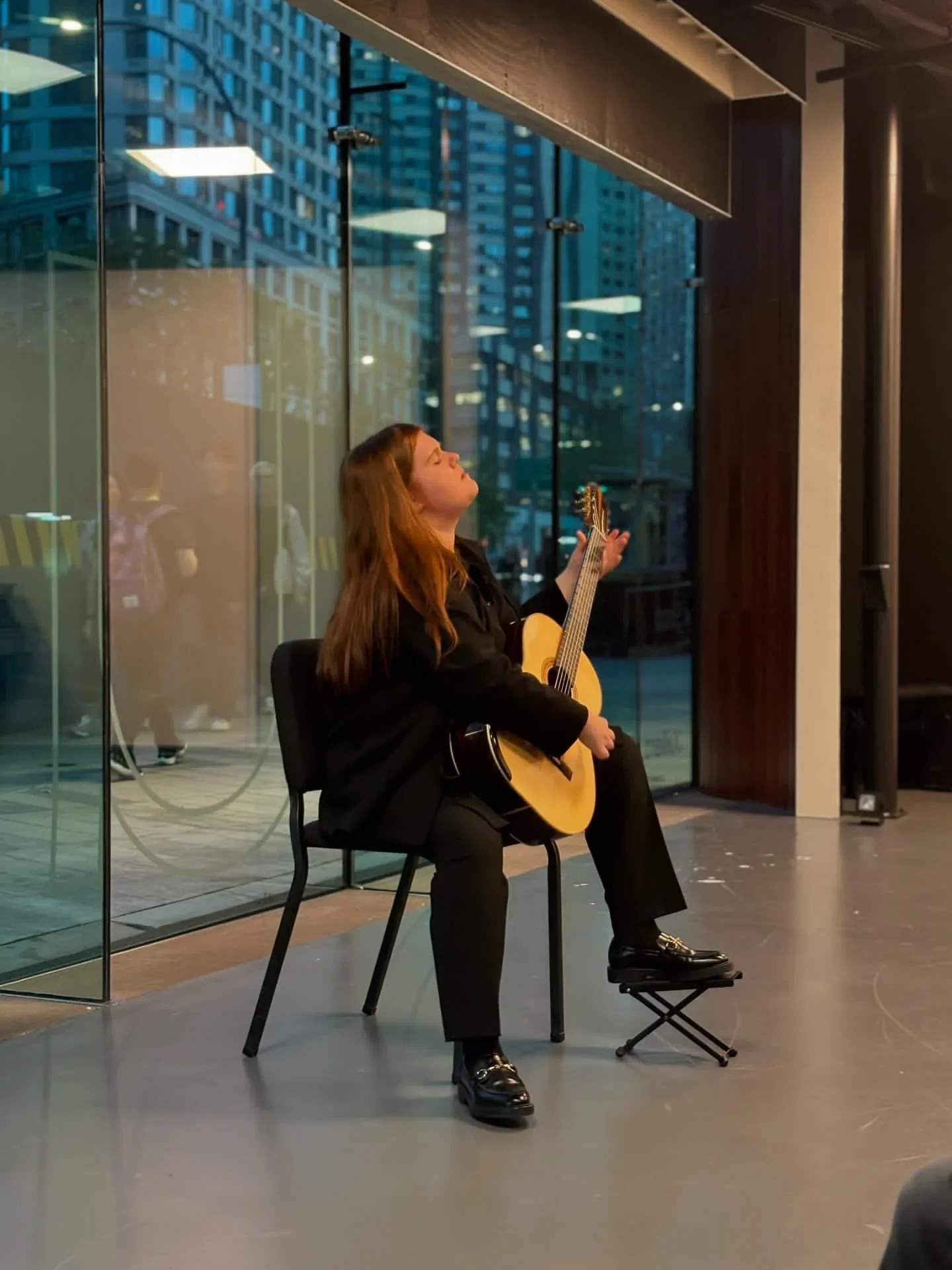 A woman playing an acoustic guitar and singing with her eyes closed, seated on a black chair inside a modern building with glass walls and high-rise city buildings outside.
