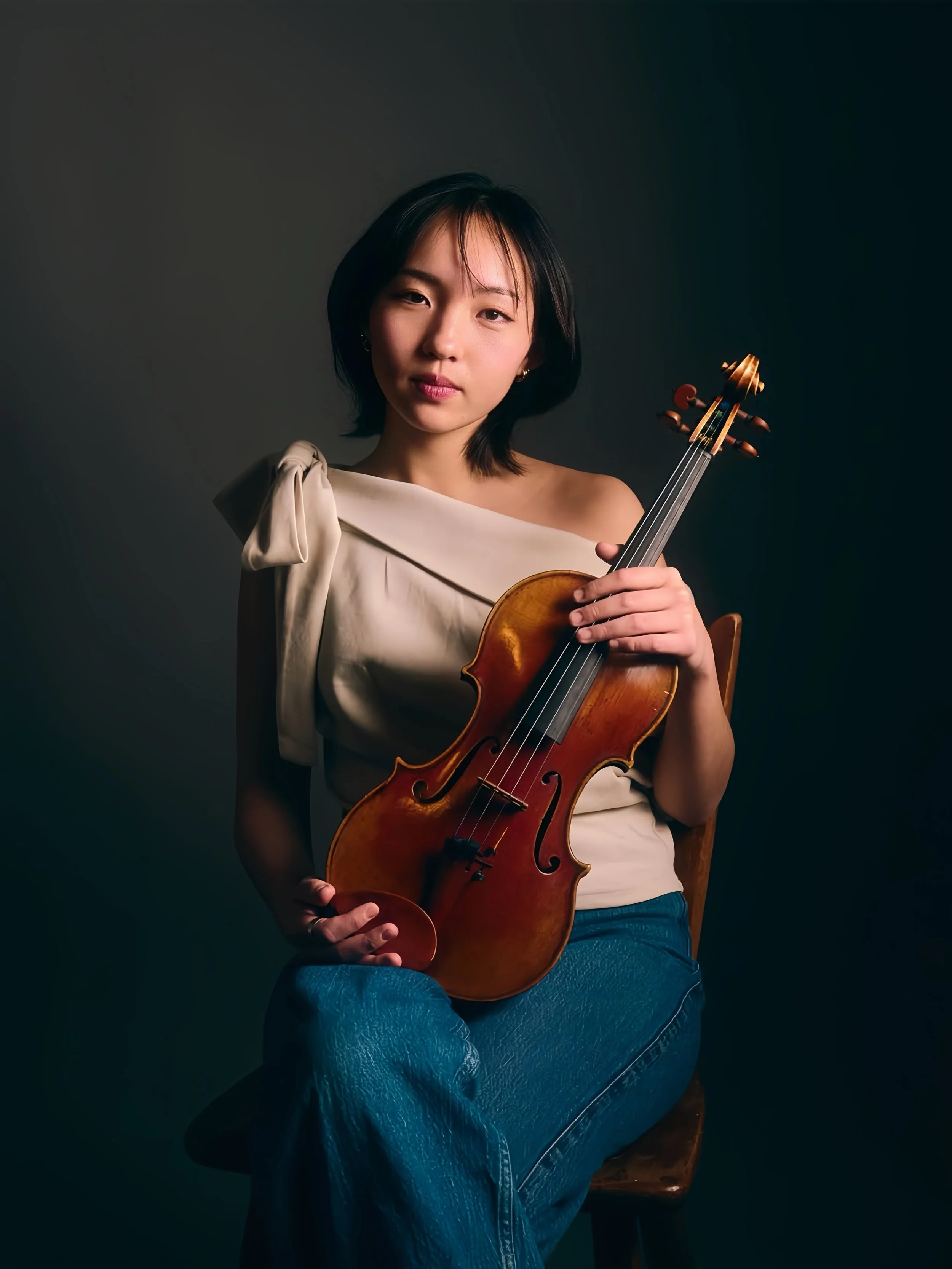 A young woman with short black hair, wearing a cream-colored top and blue jeans, sitting on a wooden chair and holding a violin.