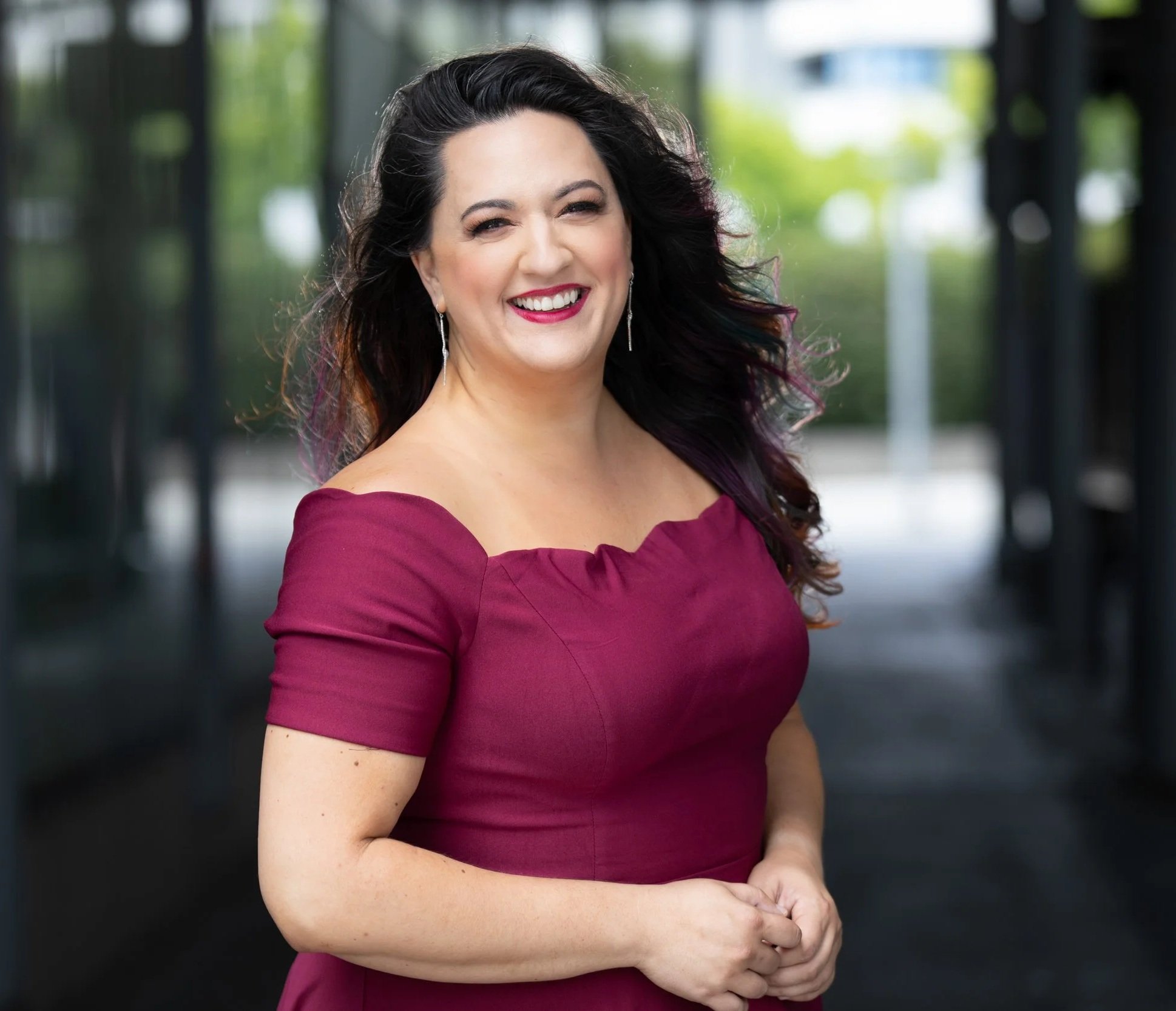 A woman with long dark hair wearing a burgundy off-the-shoulder dress, smiling outdoors with blurred background of trees and a glass building.