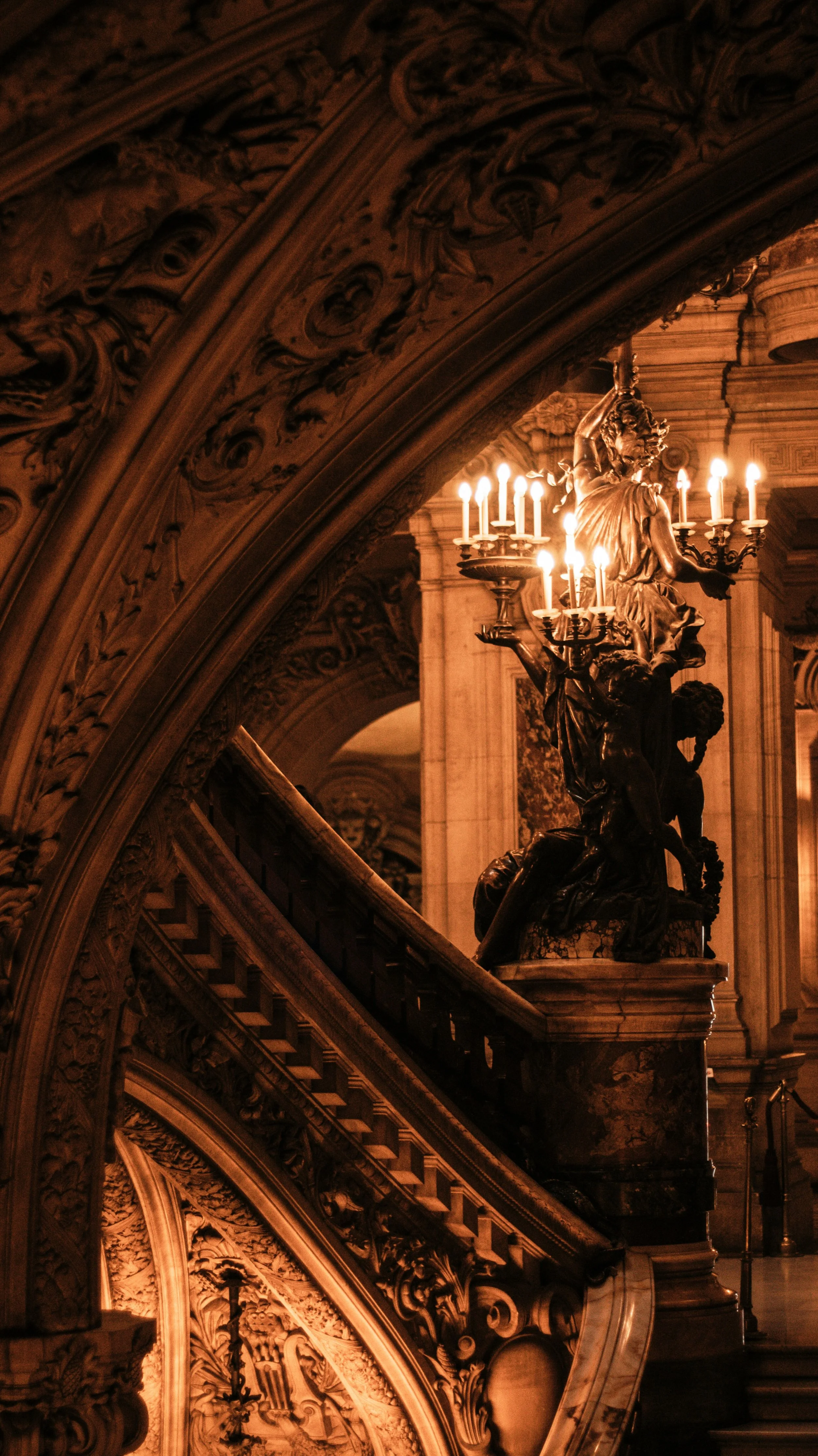 Intricate, baroque-style staircase with ornate wood carvings and a large bronze statue featuring a figure holding candles in a chandelier, inside a grand, historic building.