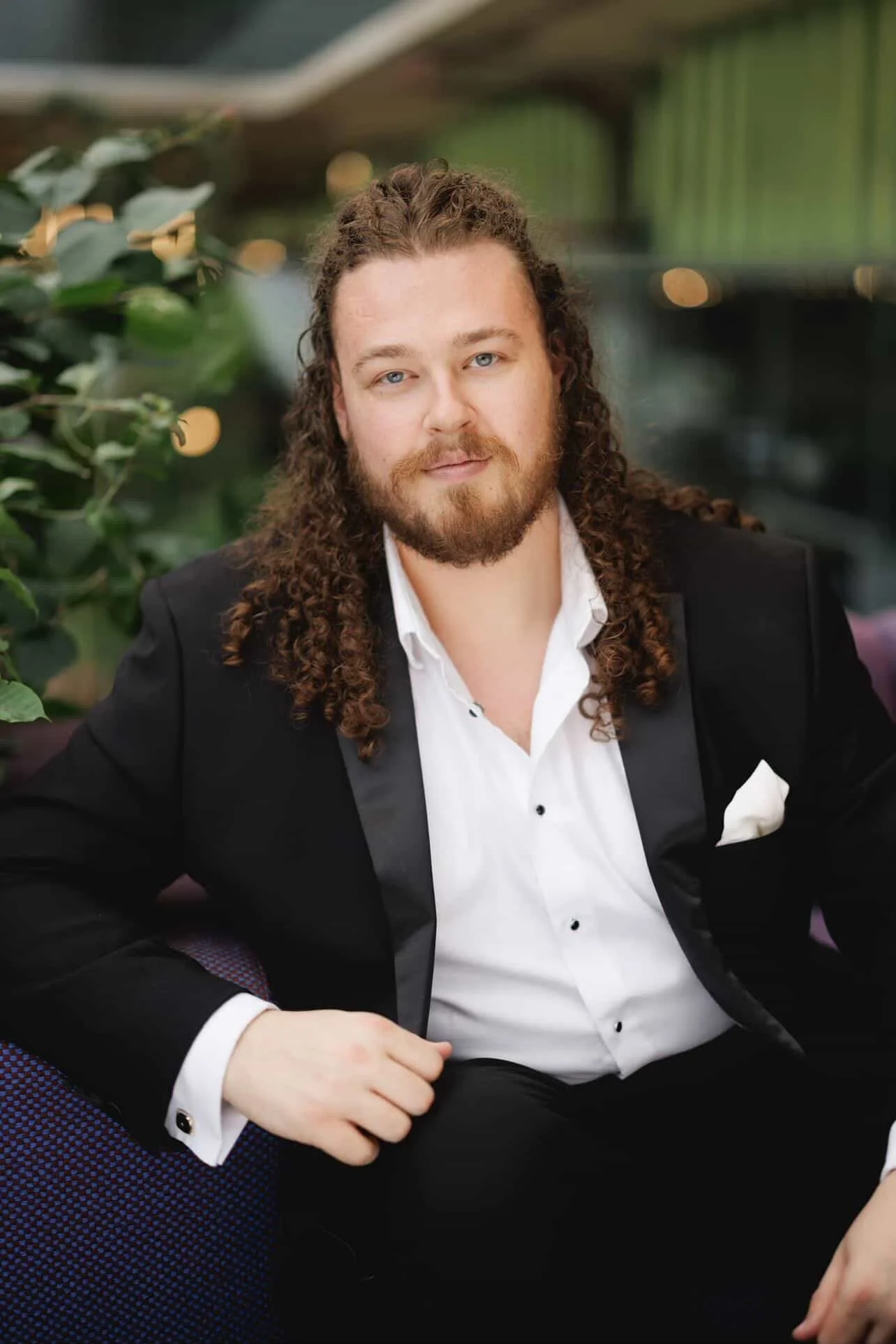 A man with long curly hair and a beard wearing a tuxedo with a white shirt, sitting on a purple chair in front of a background with green foliage.