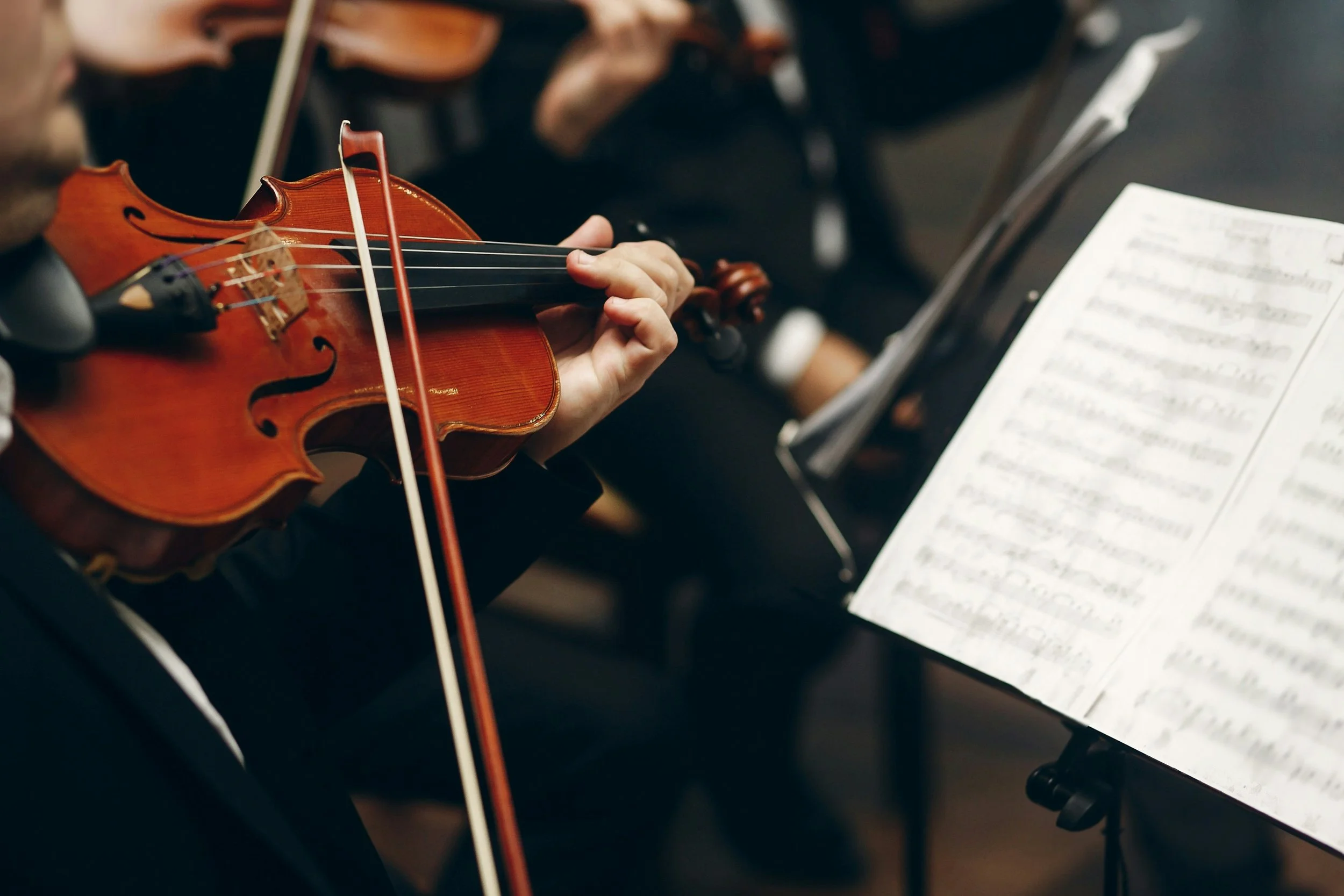 Close-up of a musician's hand playing a violin, with music sheet on a stand nearby and other musicians in the background.