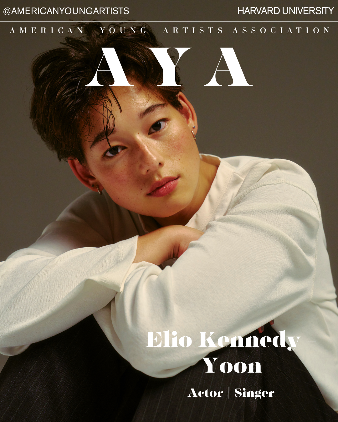 Close-up portrait of Yoon Elio Kennedy, an actor and singer, with short dark hair and earrings, resting his chin on his crossed arms, wearing a white long-sleeve shirt against a gray background.