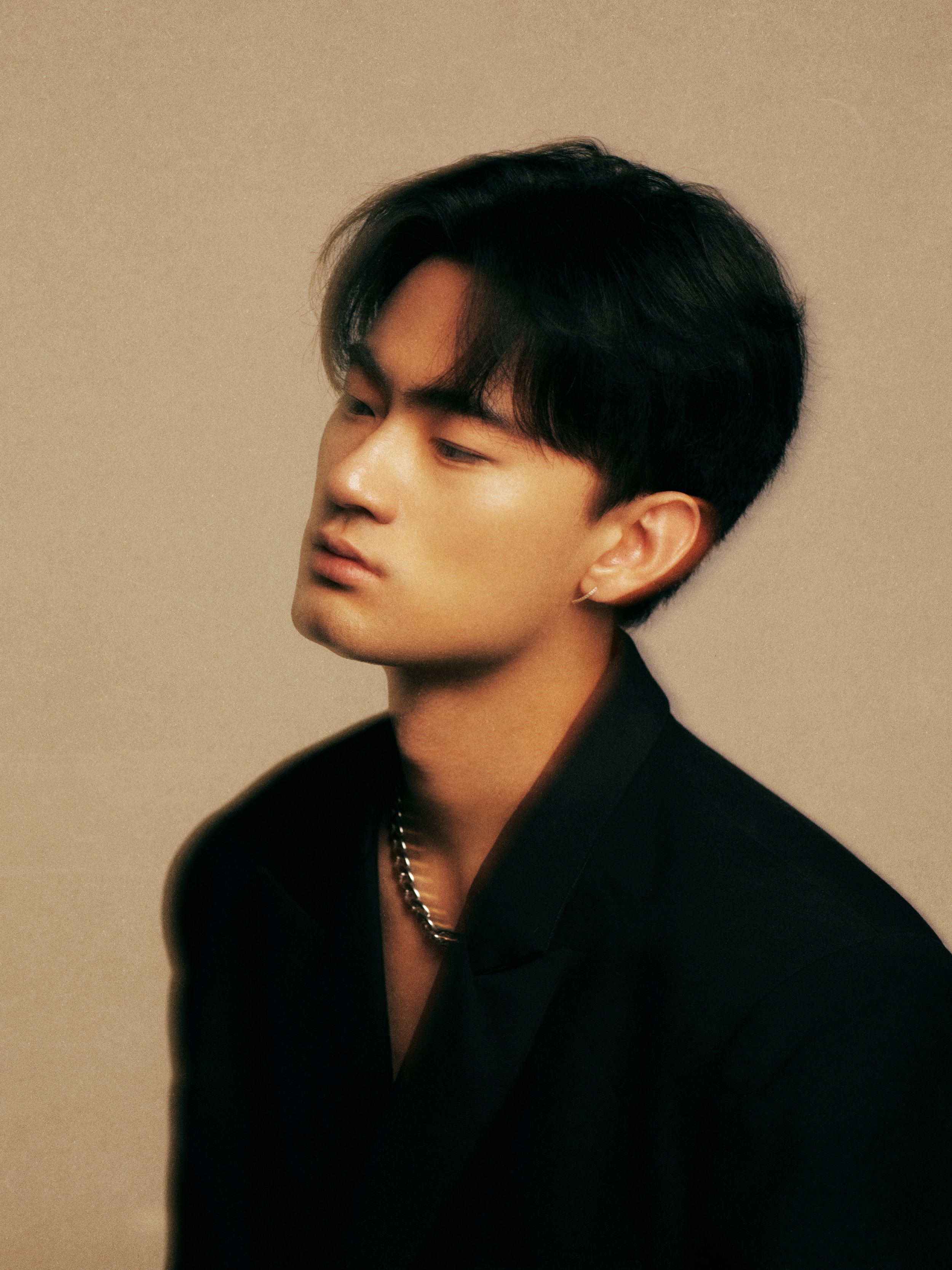 A young man with dark hair and fair skin is posing against a neutral background, wearing a black outfit and a silver chain necklace, with a contemplative expression.