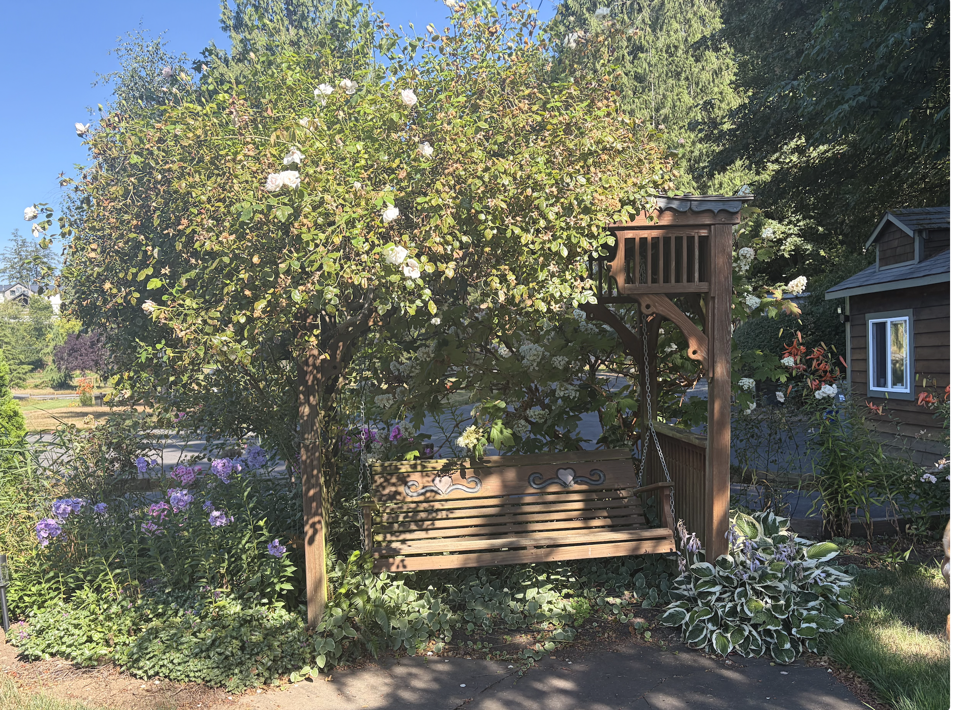 A garden scene with a wooden porch swing hanging from a wooden frame among green trees and flowering plants, including pink, white, and purple flowers, under a clear blue sky.