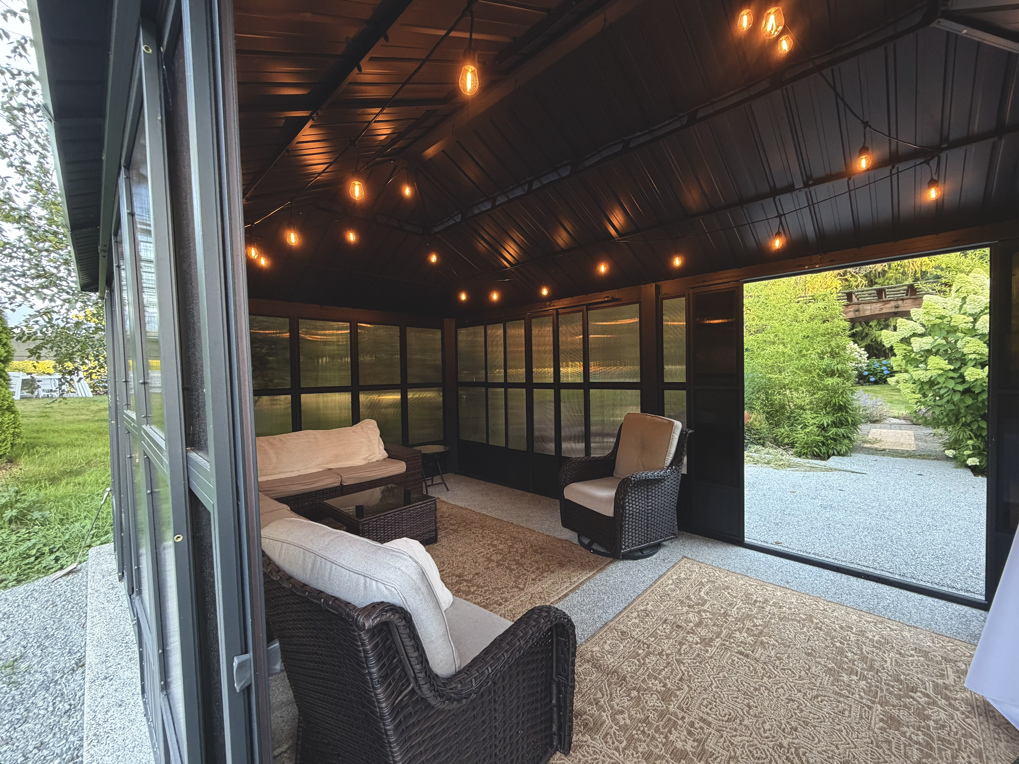 A screened-in porch area with wicker chairs, a sofa, and a coffee table. The ceiling has hanging string lights with exposed bulbs. Outside, there are lush green trees and a garden with various plants visible through the screen panels.