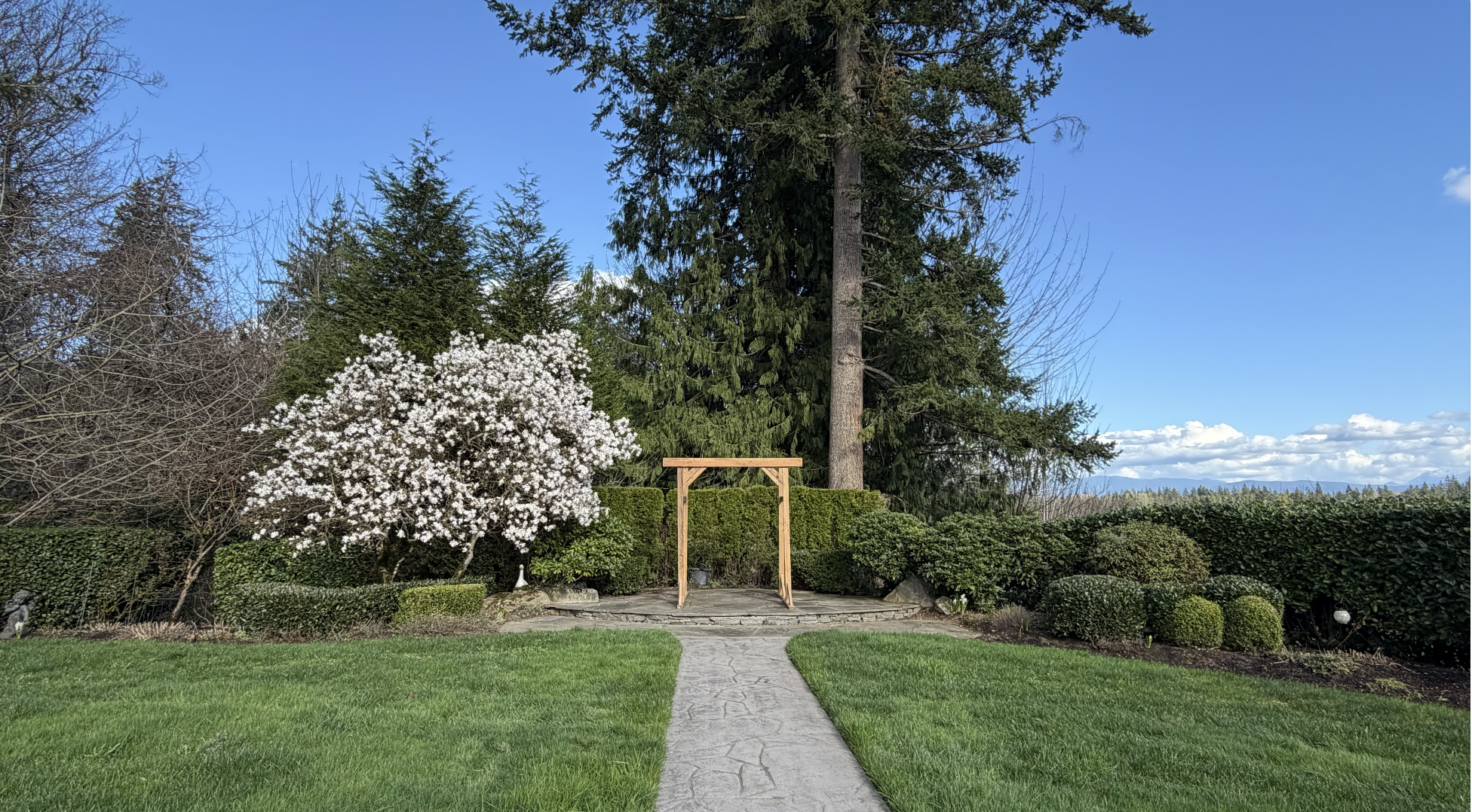 A garden scene with a stone pathway leading to a wooden arch, surrounded by trimmed bushes, a flowering tree with white blossoms, tall evergreen trees, and a blue sky with scattered clouds.
