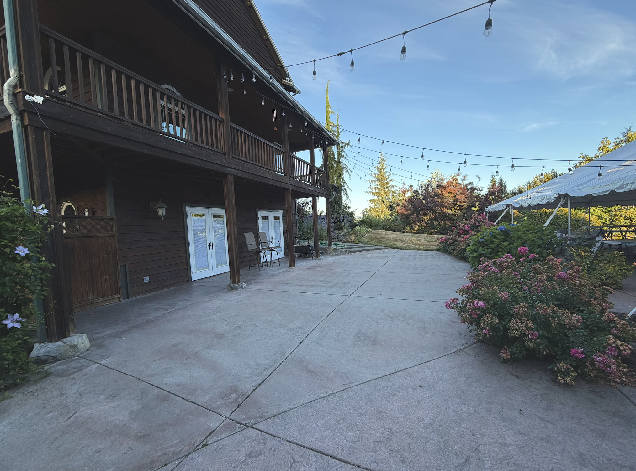 View of a backyard with a concrete patio, garden beds with pink and purple flowers, a white tent with string lights, and a two-story wooden house with a covered balcony, during daytime with clear blue sky.