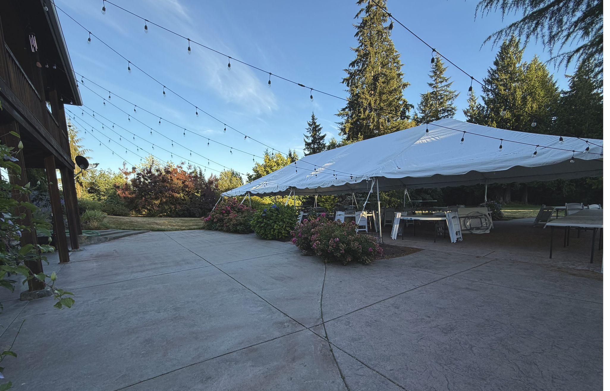 An outdoor patio area with string lights hanging overhead, a large white tent with tables and chairs underneath, surrounded by colorful flowers and trees on a sunny day.