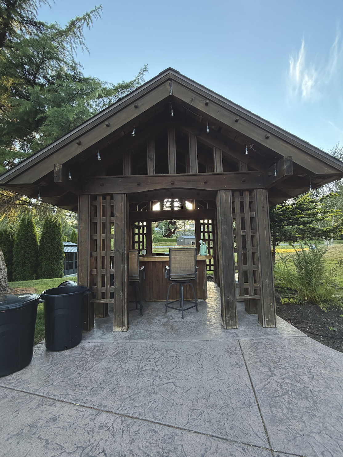 A wooden outdoor bar pavilion with string lights, surrounded by greenery, with two trash cans on the left and two chairs at the bar counter.