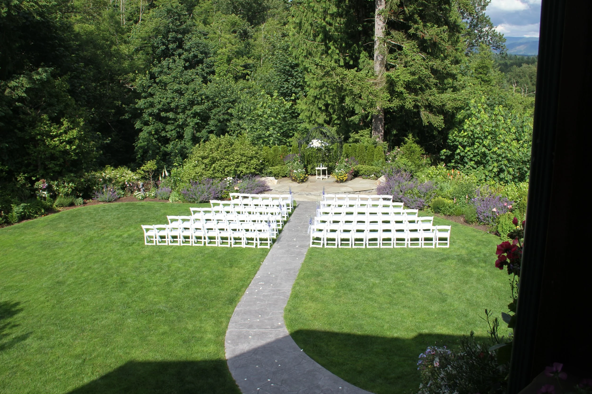 Outdoor wedding setup with white chairs arranged in rows on a green lawn facing an altar with a small table, surrounded by lush trees and colorful flowers, with a clear sky in the background.