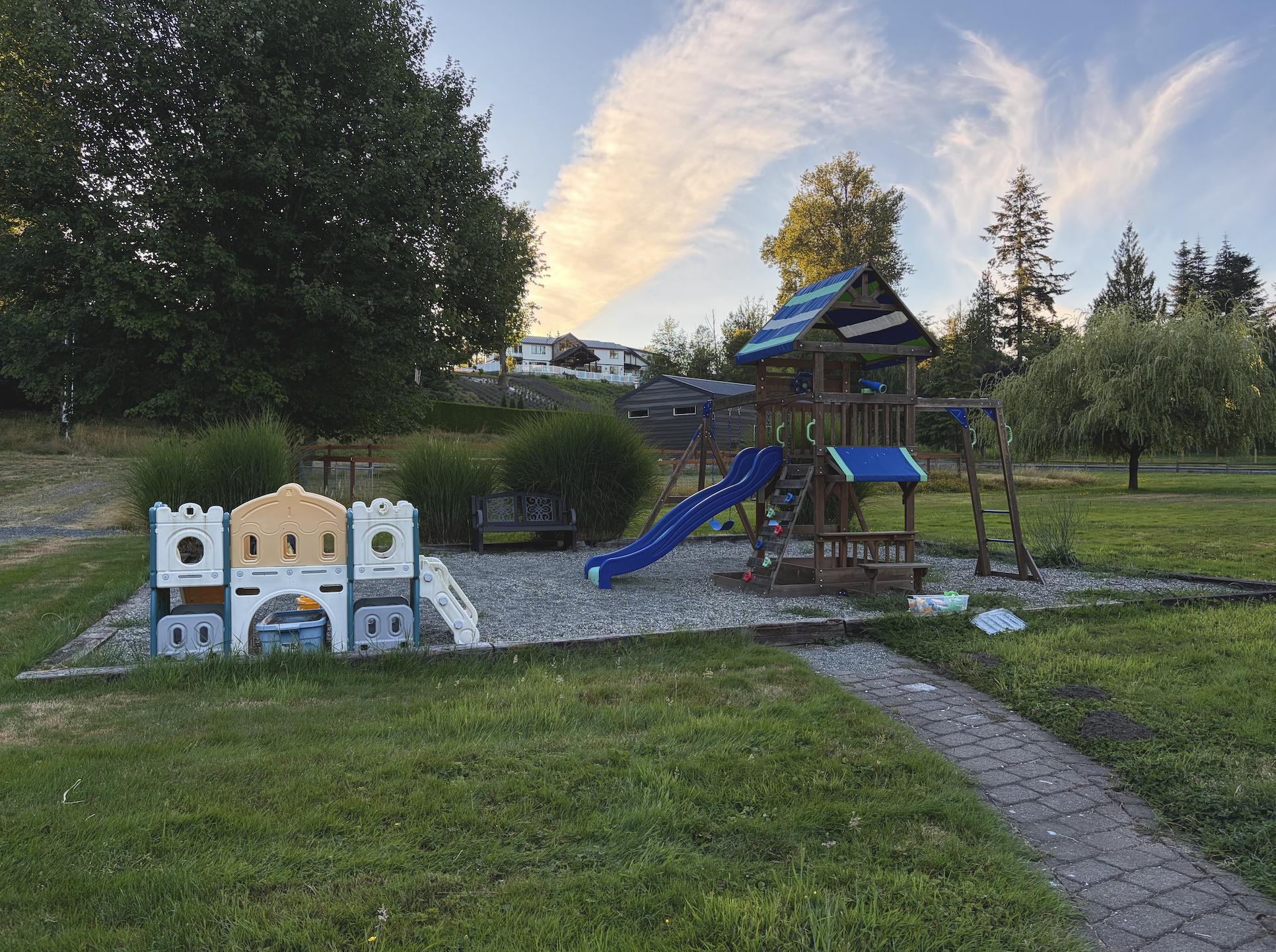 A children's playground with a wood and blue slide, sandbox, small playhouse, and swings, set on a gravel surface in a grassy yard, with trees and houses in the background during sunset.