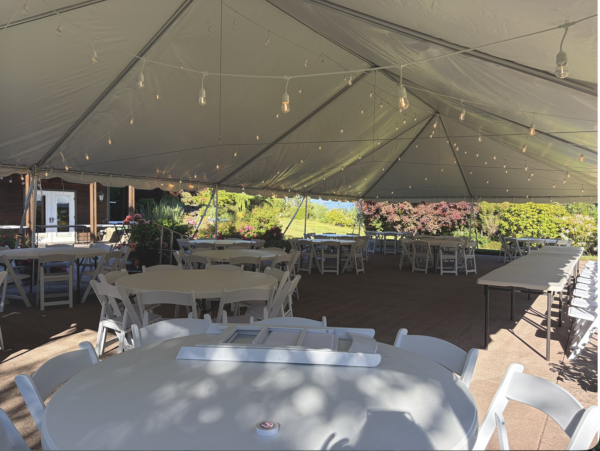 Under a large outdoor tent, white circular tables and chairs are arranged for an event. String lights hang from the tent ceiling, and there are flowers and greenery in the background.