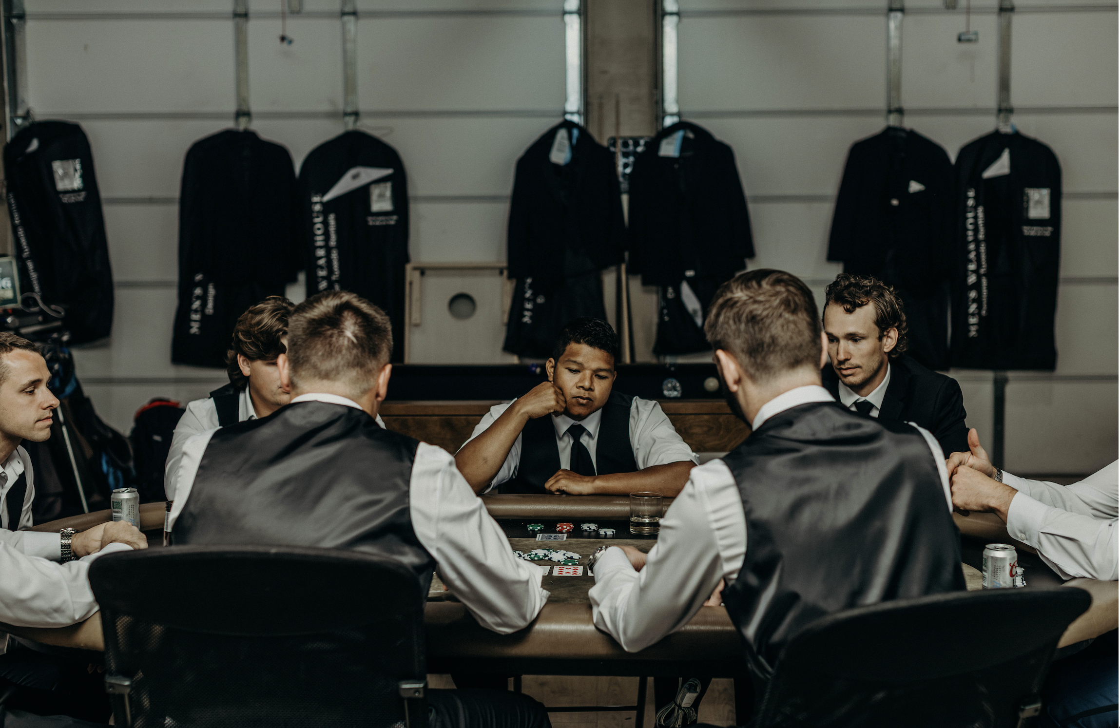 A group of young men in formal attire playing poker around a table in a garage with poker chips, drinks, and a deck of cards, with black jackets hanging on the wall in the background.